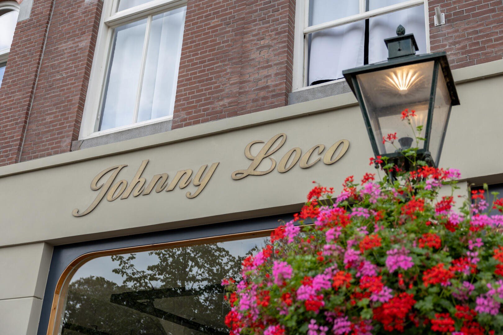 Eine Gebäudefassade mit dem Schriftzug Johnny Loco in kursiven Buchstaben, eine Lampe im Vintage-Stil und ein hängender Korb mit leuchtend rosa und roten Blumen im Vordergrund.