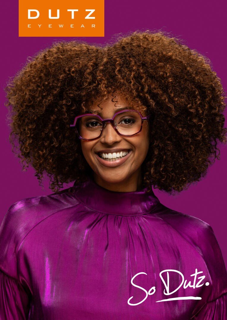 Eine lächelnde Frau mit lockigem Haar trägt eine lila Brille und ein glänzendes lila Oberteil und steht vor einem lila Hintergrund. Auf dem Bild ist Dutz Eyewear und So Dutz. zu lesen.