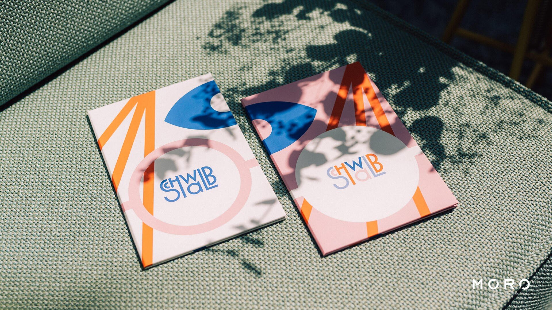 Zwei bunte Karten mit abstrakten Designs und dem Text SHWB und SHWBLAB liegen im Sonnenlicht auf einer strukturierten grünen Oberfläche, wobei Pflanzenschatten und MORO in der unteren rechten Ecke sichtbar sind.