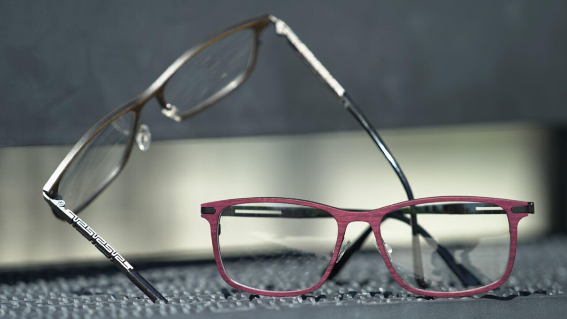 Two pairs of eyeglasses are displayed: one with a metallic frame in the background, and another with a red and black frame in the foreground, both resting on a textured surface.