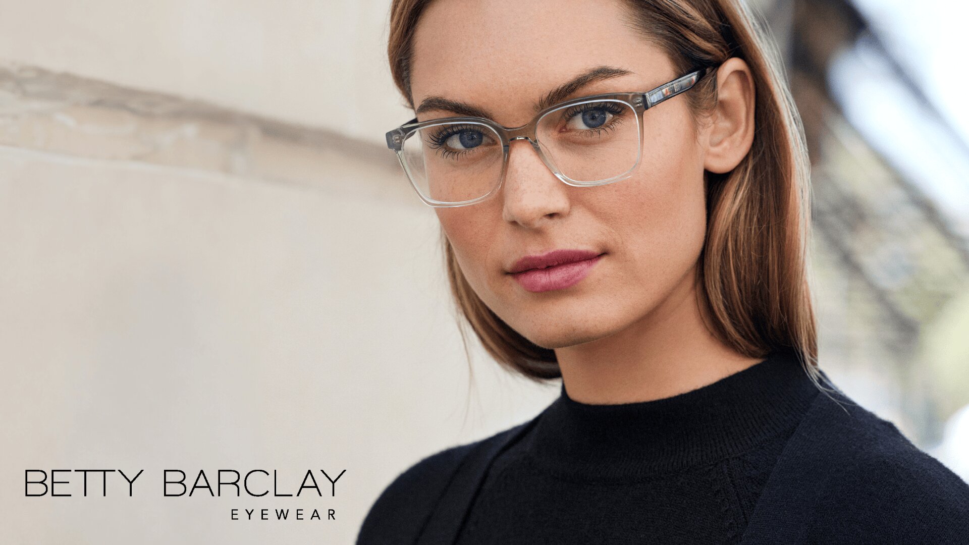 Eine Frau mit hellbraunem Haar, die eine klare, quadratische Brille und einen schwarzen Pullover trägt, blickt selbstbewusst in die Kamera. Der Text Betty Barclay Eyewear erscheint in der unteren linken Ecke.