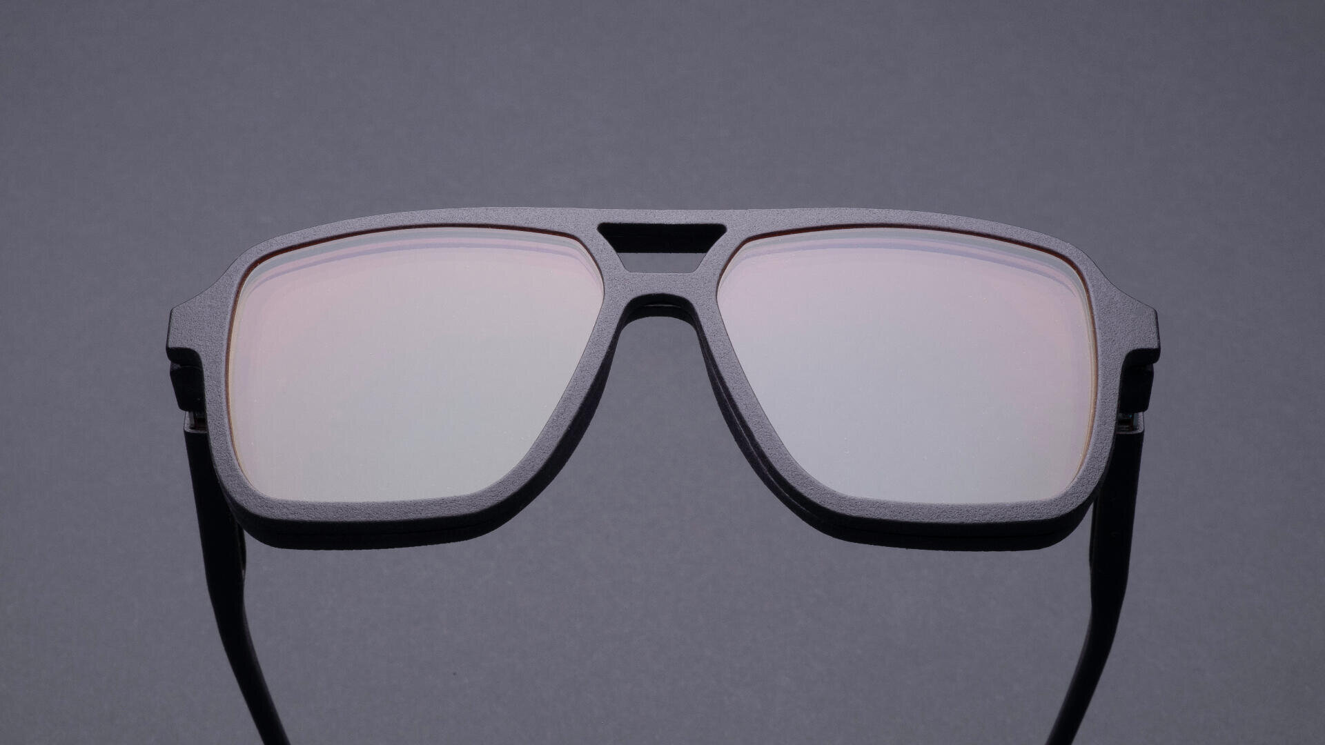 Eine moderne, mattschwarze Sonnenbrille mit großen, rechteckigen Gläsern und dickem Rahmen, fotografiert vor einem schlichten grauen Hintergrund.