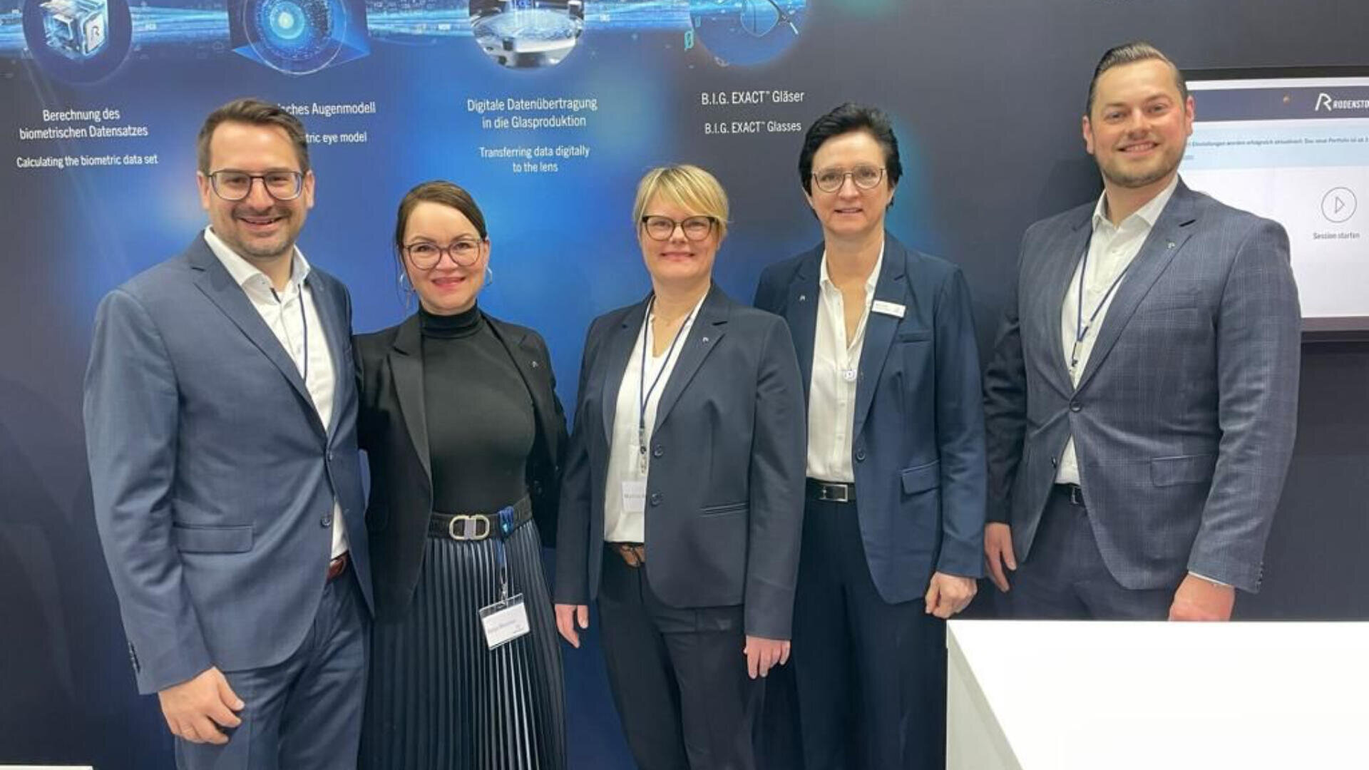 Fünf Personen in Geschäftskleidung stehen nebeneinander und lächeln in die Kamera vor einem dunkelblauen Messestand mit Grafiken und Texten zum Thema Technologie.