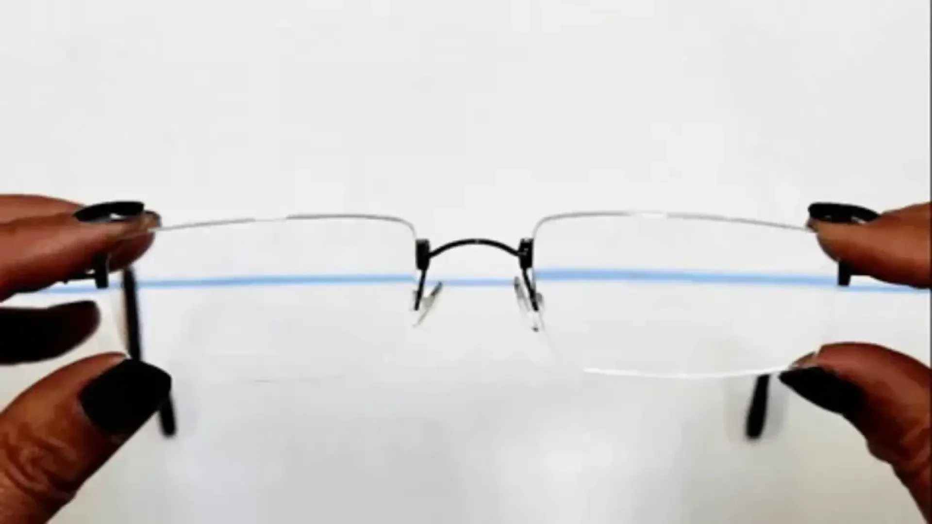 A pair of rimless eyeglasses held by two hands with dark nail polish, positioned against a white background with a blue horizontal line visible through the lenses.