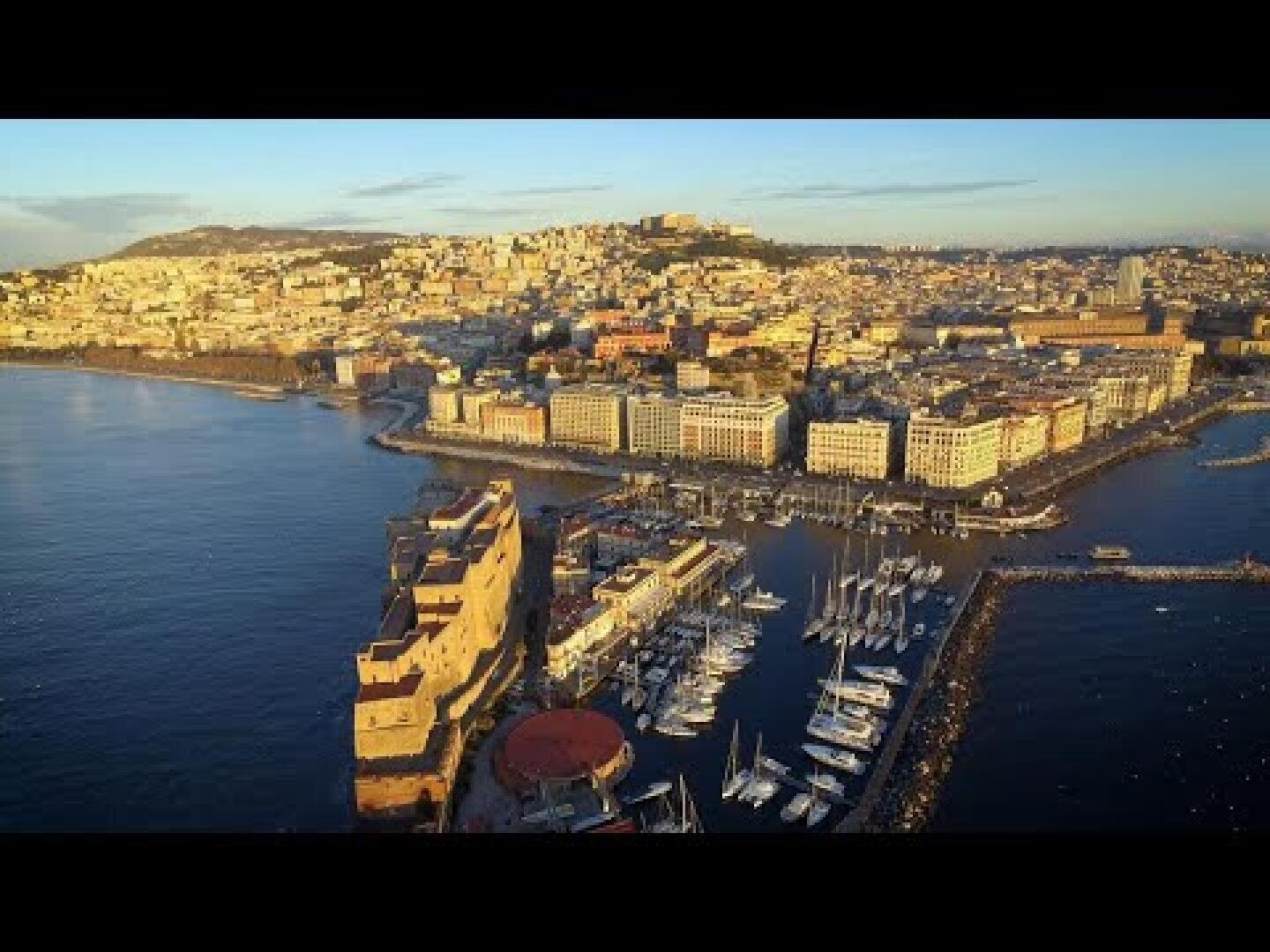 Luftaufnahme von Neapel, Italien, mit einem Yachthafen mit angedockten Booten, historischen Gebäuden entlang der Uferpromenade und der Stadt, die sich unter einem klaren Himmel bei Sonnenuntergang über die Hügel erstreckt.