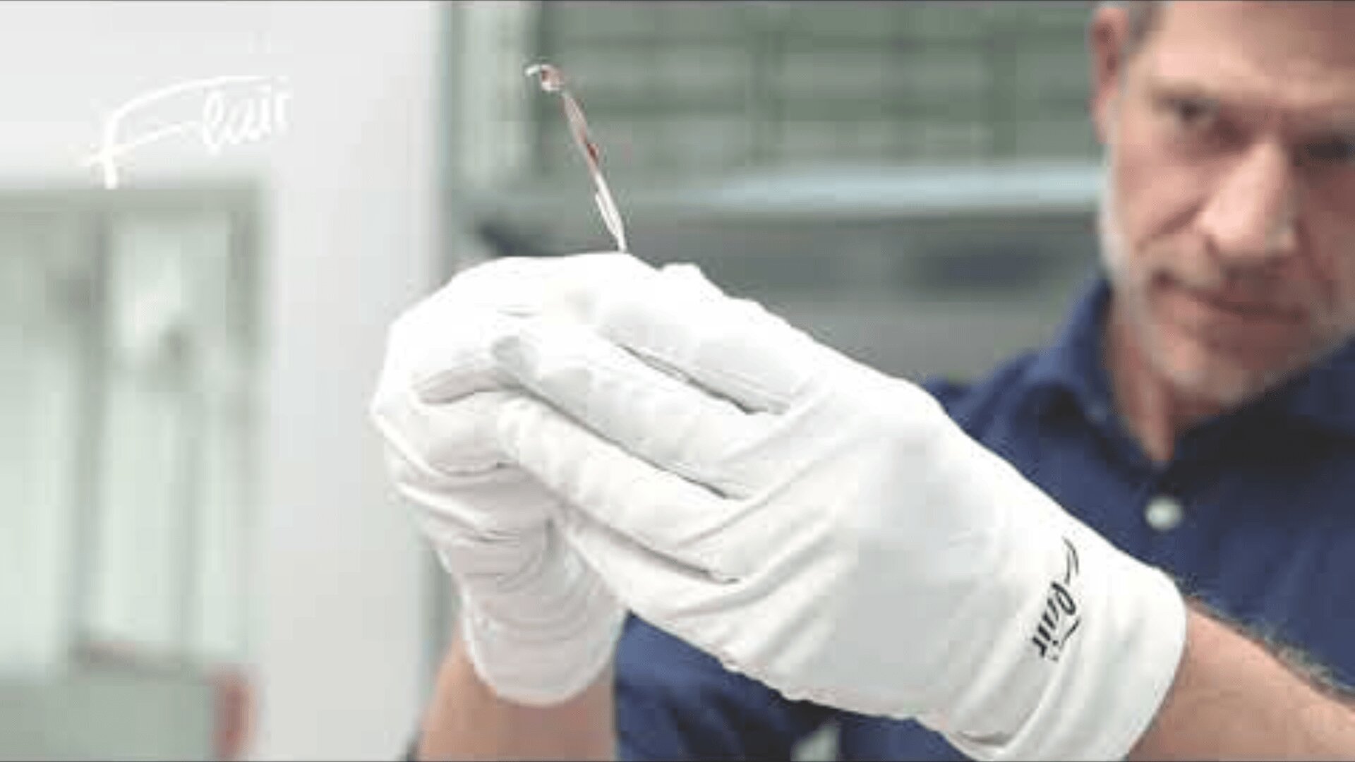 Ein Mann mit weißen Handschuhen hält vorsichtig ein dünnes, transparentes Objekt mit einer kleinen roten Spitze und untersucht es in einem Labor oder einer Werkstatt genau.