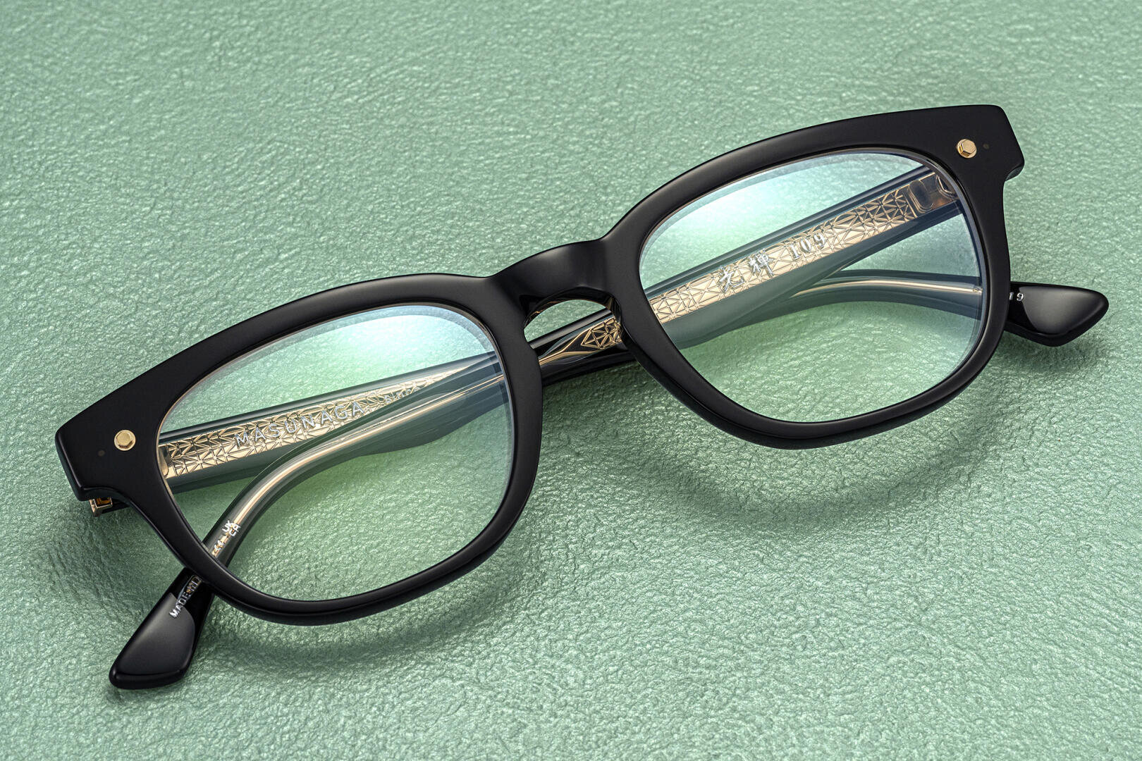 A pair of black-rimmed eyeglasses with gold decorative detailing on the arms, placed on a textured green surface.