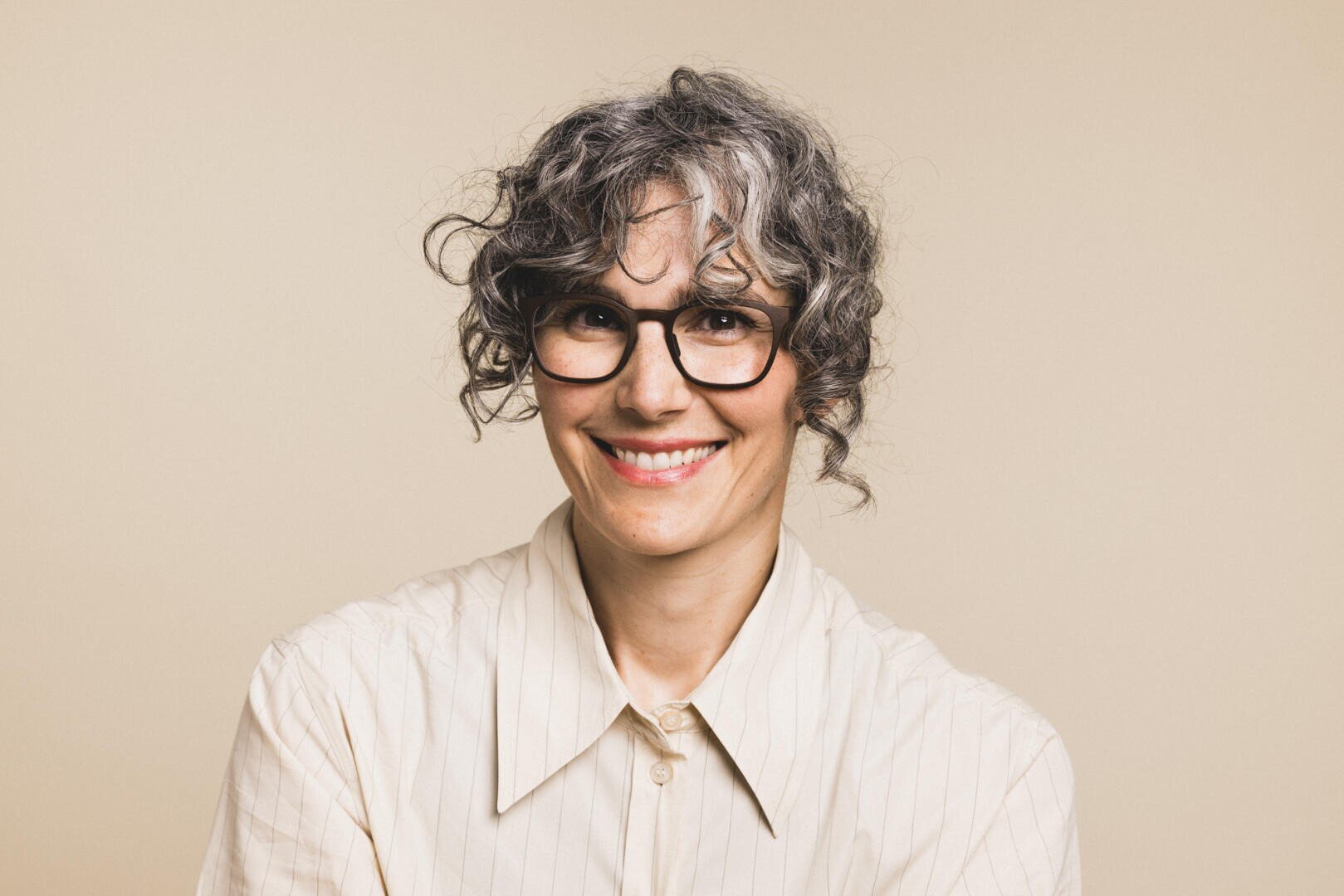 Eine lächelnde Person mit lockigem, grauem Haar und schwarzer Brille, die ein cremefarbenes Hemd mit Kragen trägt, posiert vor einem schlichten beigen Hintergrund.