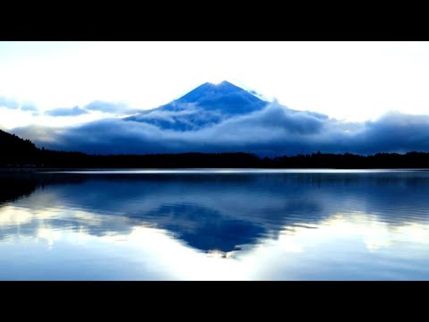 In einem ruhigen See spiegelt sich ein schneebedeckter Berg, der teilweise von Wolken verhüllt ist, mit Bäumen als Silhouette am Horizont und ruhigen Blau- und Weißtönen in der Szene.