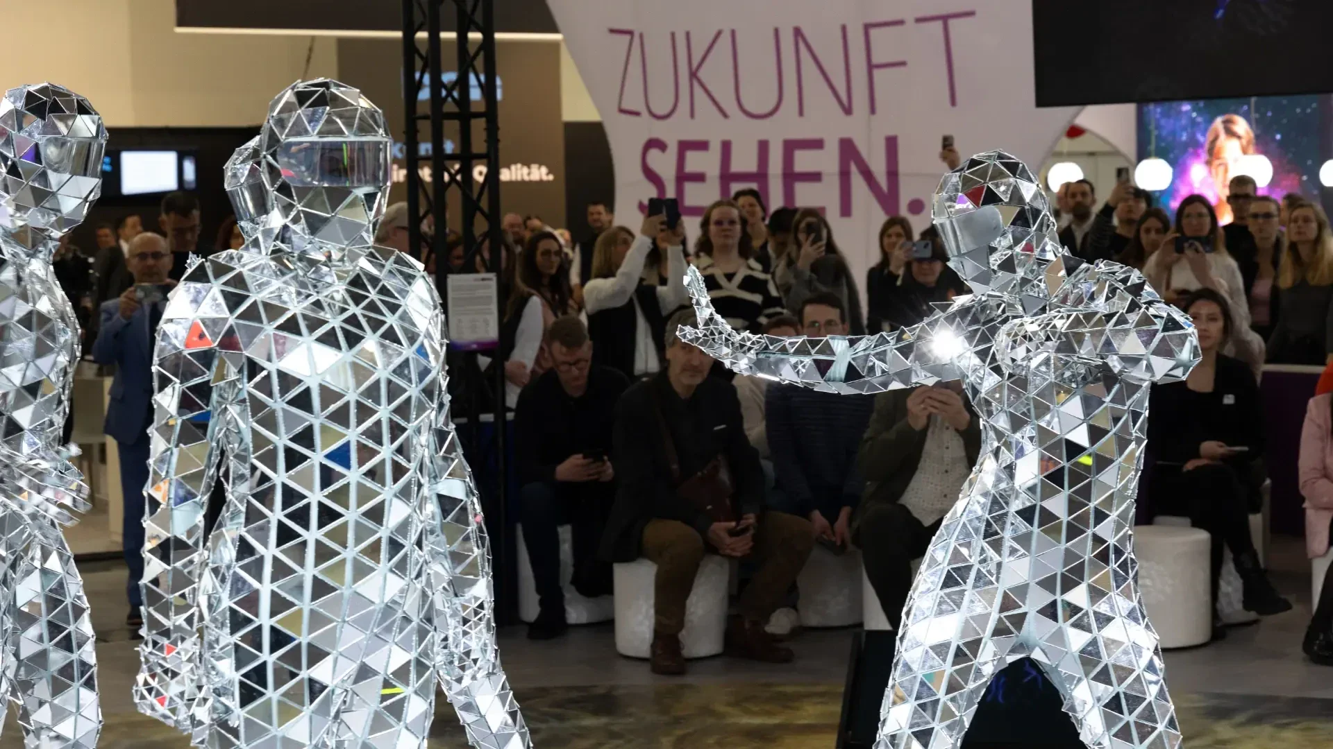 Two performers in full-body mirrored suits pose in front of a crowd at an indoor event. The background features a sign with large German text reading Zukunft sehen. Spectators watch and take photos.