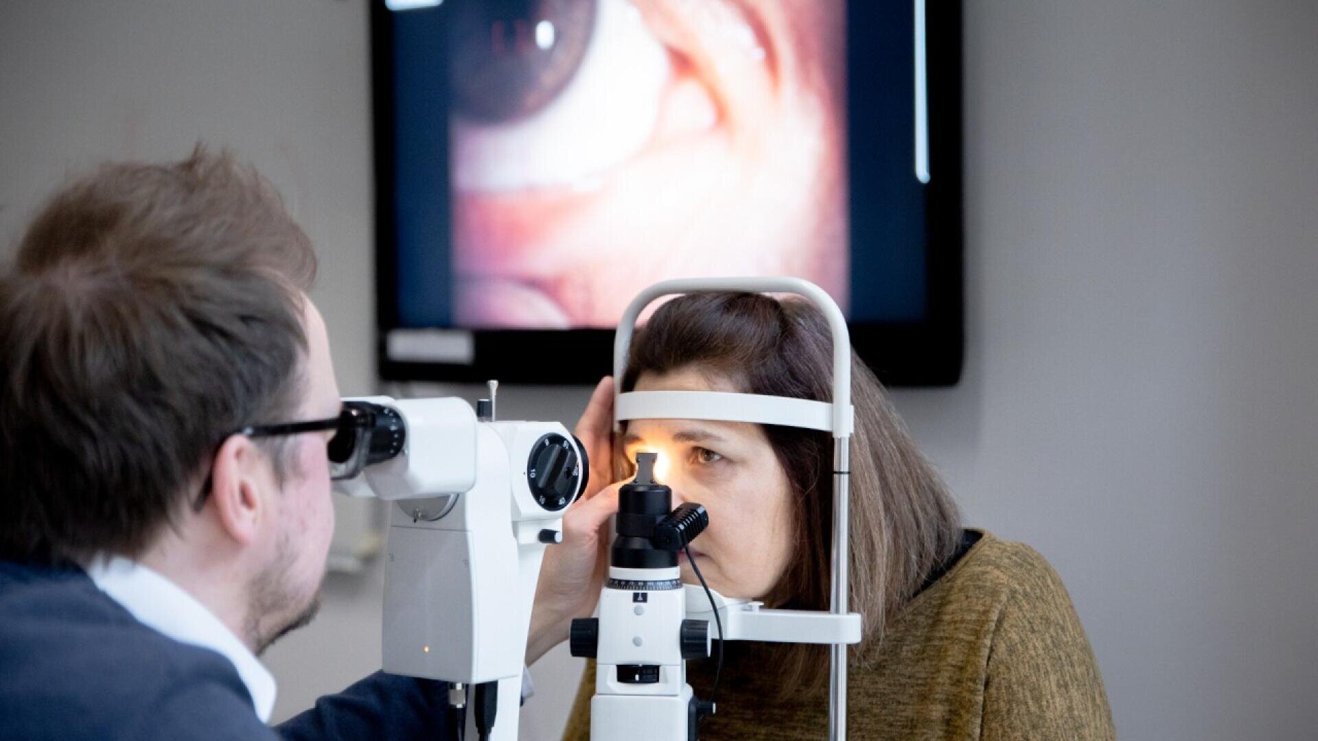 An optometrist examines a womans eye using a slit lamp, while a close-up image of an eye is displayed on a screen in the background.