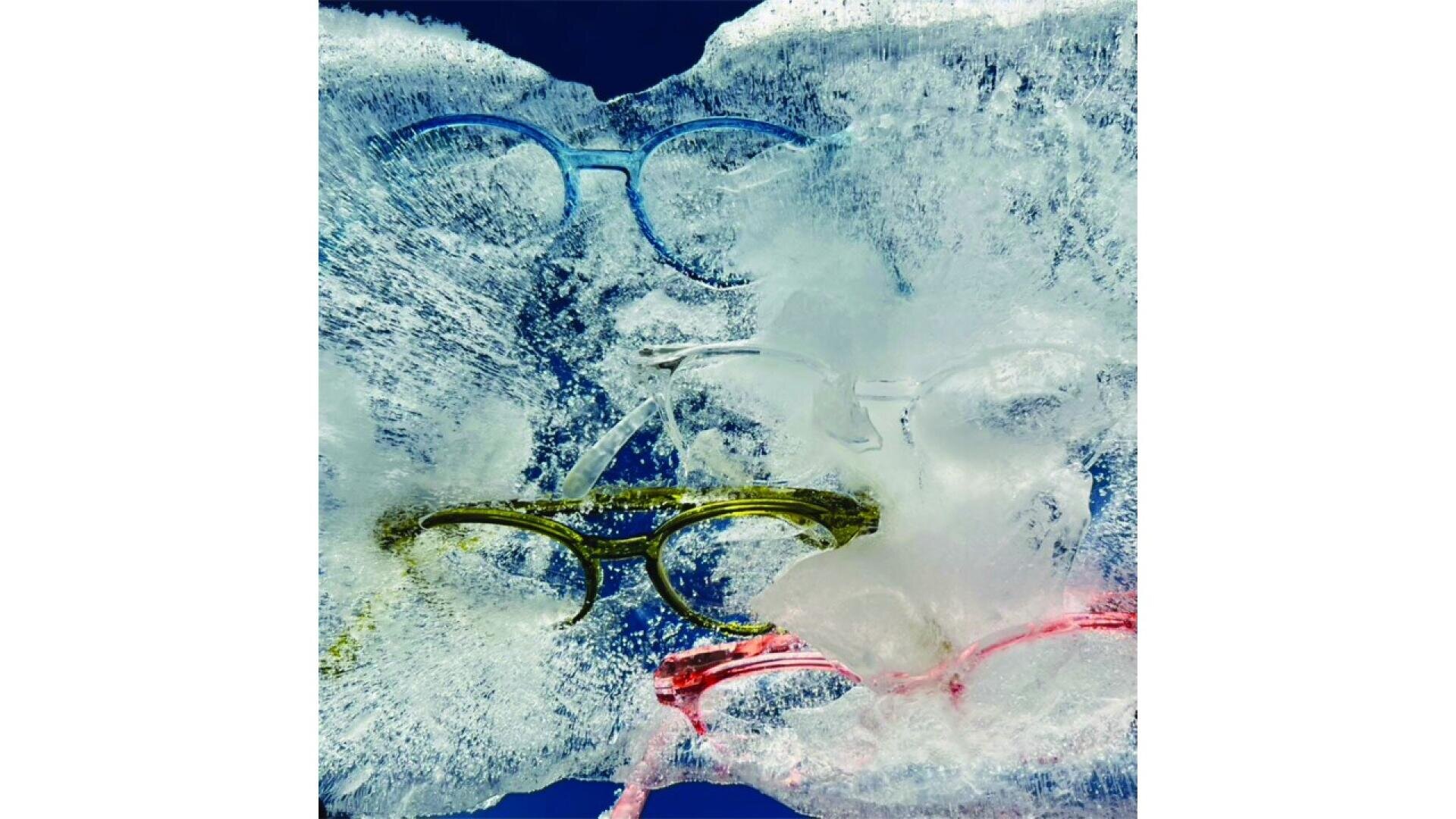 Vier bunte Brillen – blau, transparent, gelb und rosa – sind in einem dicken Eisblock vor einem dunkelblauen Hintergrund eingefroren. Die Brillen sind teilweise durch das Eis sichtbar.