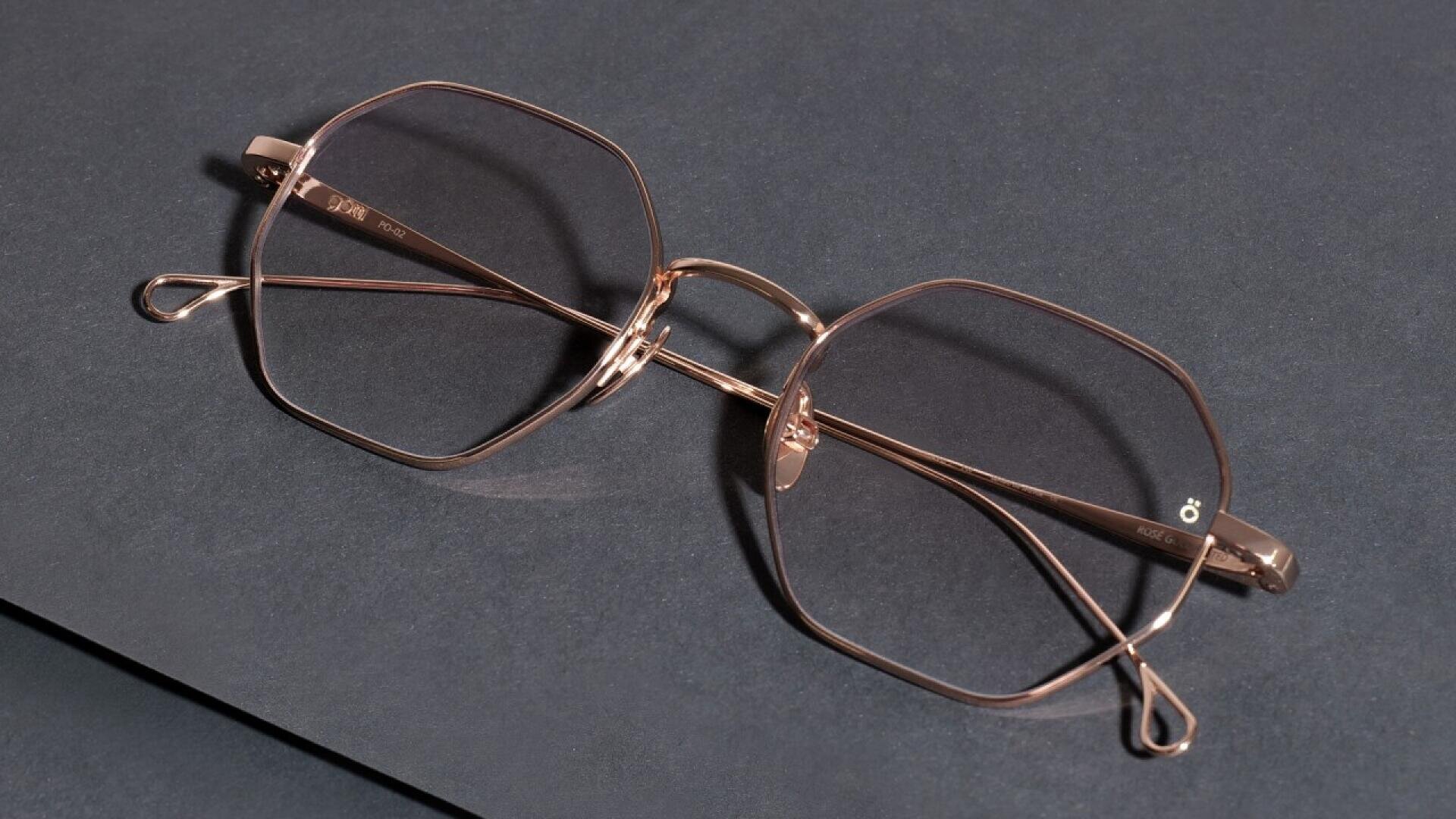 A pair of rose gold, geometric eyeglasses with thin metal frames is placed on a dark, textured surface.