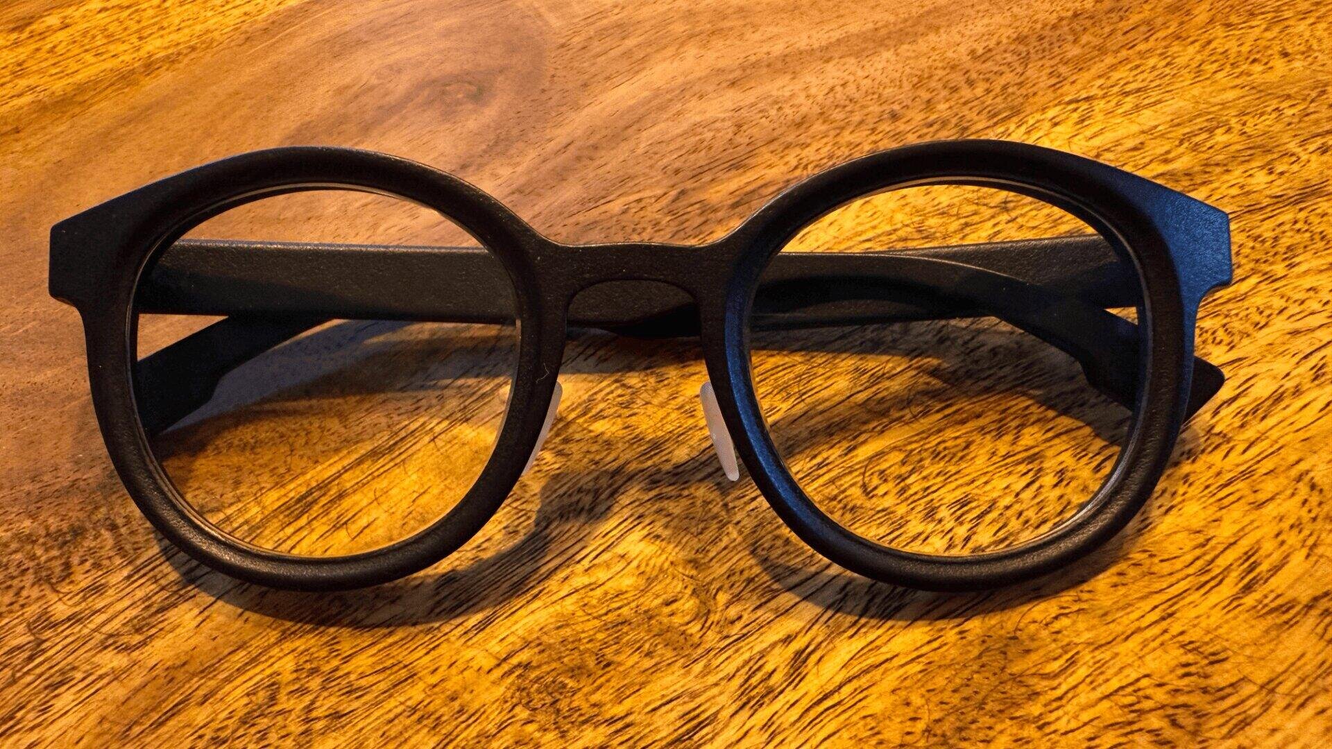 Eine runde, schwarz umrandete Brille mit dickem Rahmen ruht auf einer Holzoberfläche mit warmer, brauner Maserung.