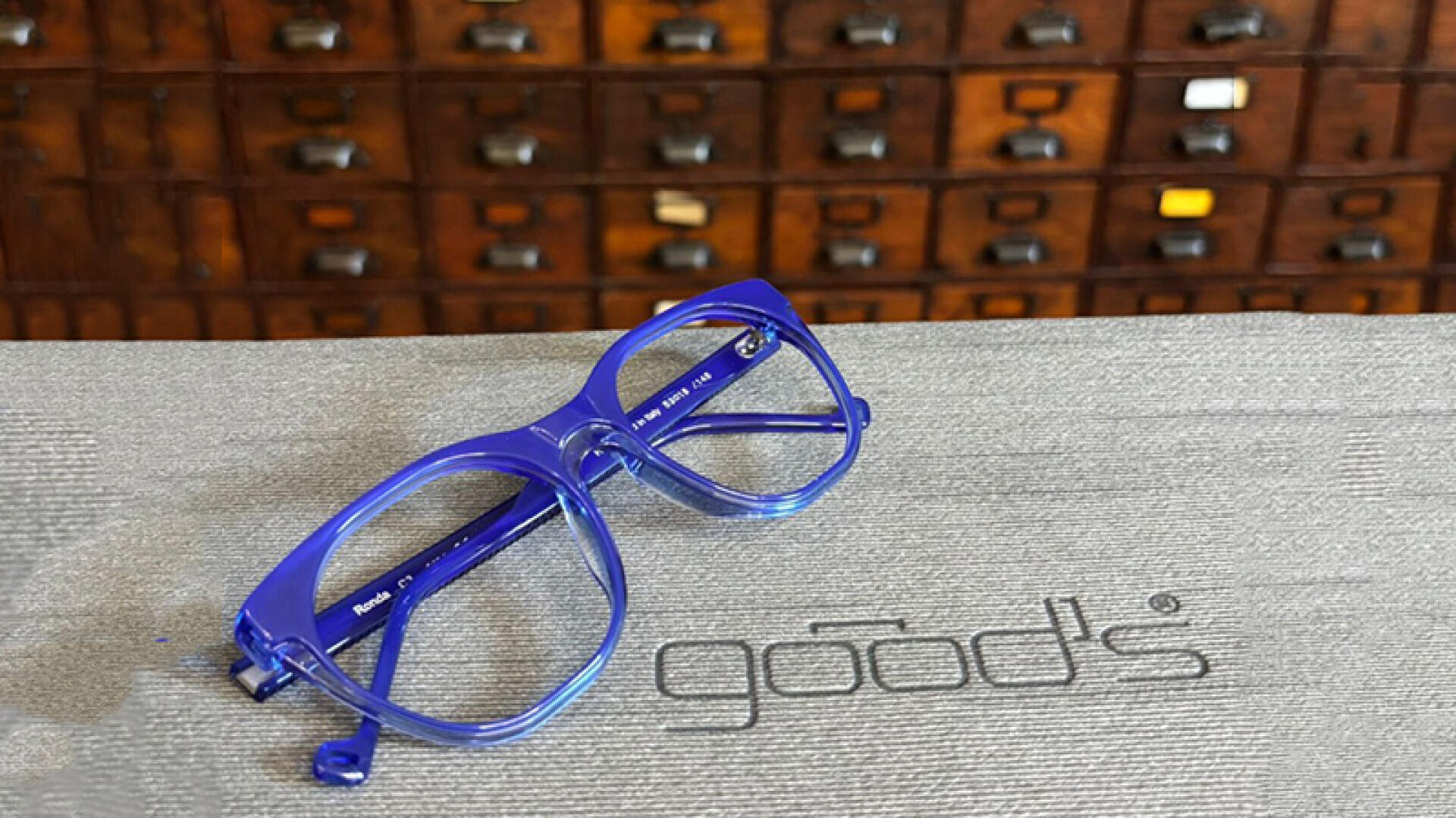A pair of light blue glasses lies on a textured gray surface with the word "Ware" printed on it. In the background is a wooden cabinet with many small drawers.