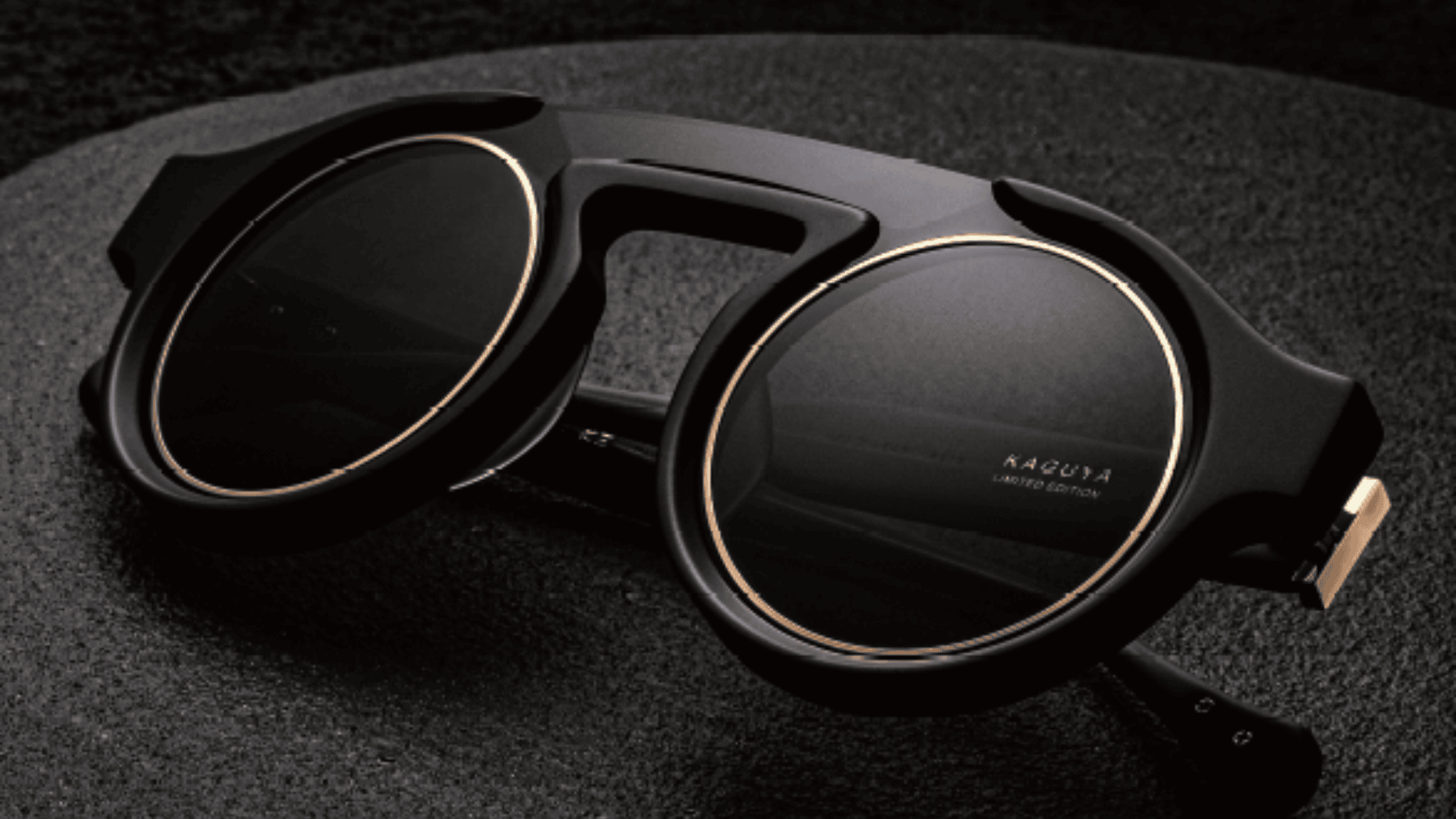 Nahaufnahme einer stilvollen, runden schwarzen Sonnenbrille mit goldenen Akzenten auf einer dunklen, strukturierten Oberfläche. Der Rahmen ist markant und modern, und auf einem Glas ist der Schriftzug „KAGUYA LIMITED EDITION“ eingraviert.