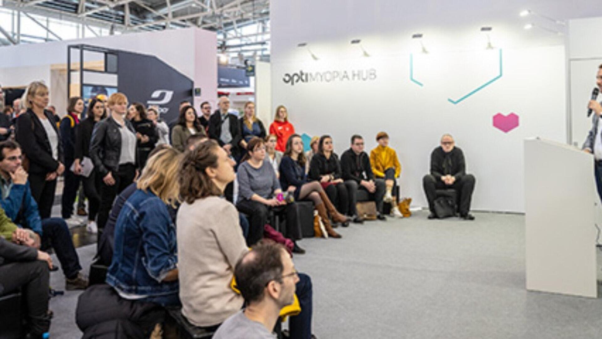 A group of people sits and listens attentively to a speaker at a modern indoor event space labeled MORPH HUB. The audience faces the stage, and the atmosphere appears professional and focused.