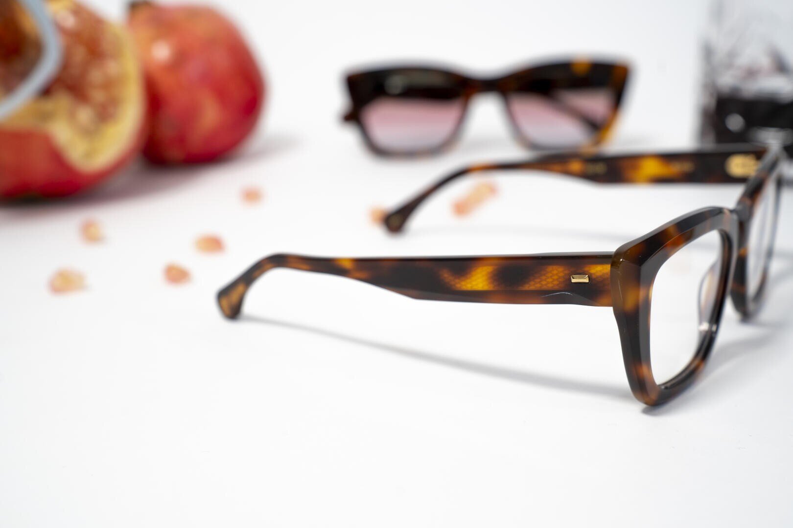 Eine Nahaufnahme einer Schildpattbrille auf einer weißen Fläche, mit einer weiteren Sonnenbrille, verstreuten kleinen pfirsichfarbenen Perlen und verschwommenen roten Äpfeln im Hintergrund.
