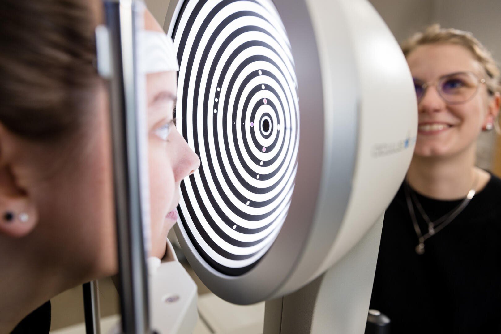 Eine Person sitzt mit dem Gesicht in der Nähe einer Augenuntersuchungsmaschine, die konzentrische schwarze und weiße Kreise anzeigt; eine andere Person, möglicherweise ein Techniker, beobachtet und lächelt im Hintergrund.