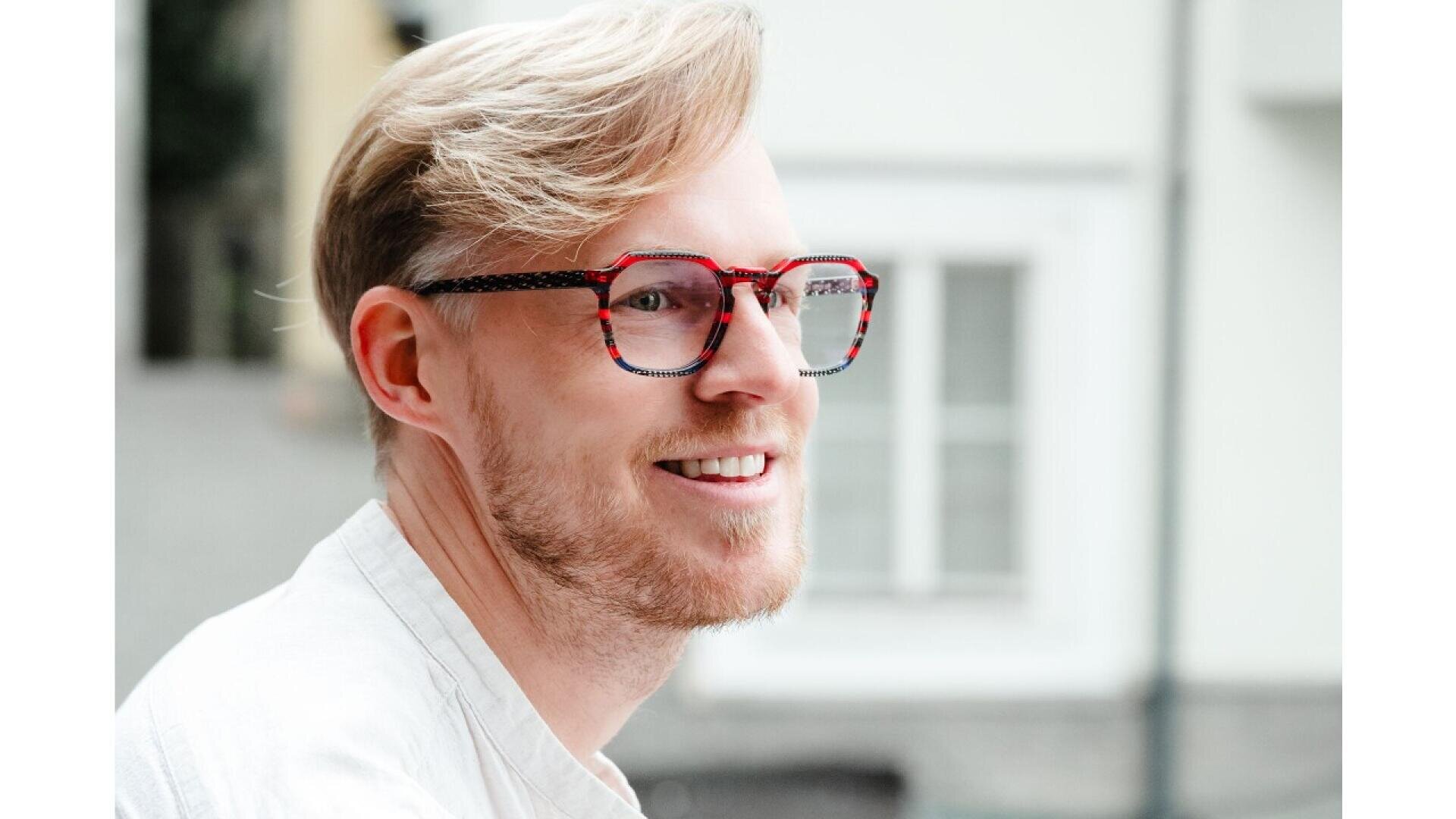 A smiling man with light blond hair and a short beard is wearing large, red-framed glasses and a white shirt, standing outdoors in front of a blurred building background.