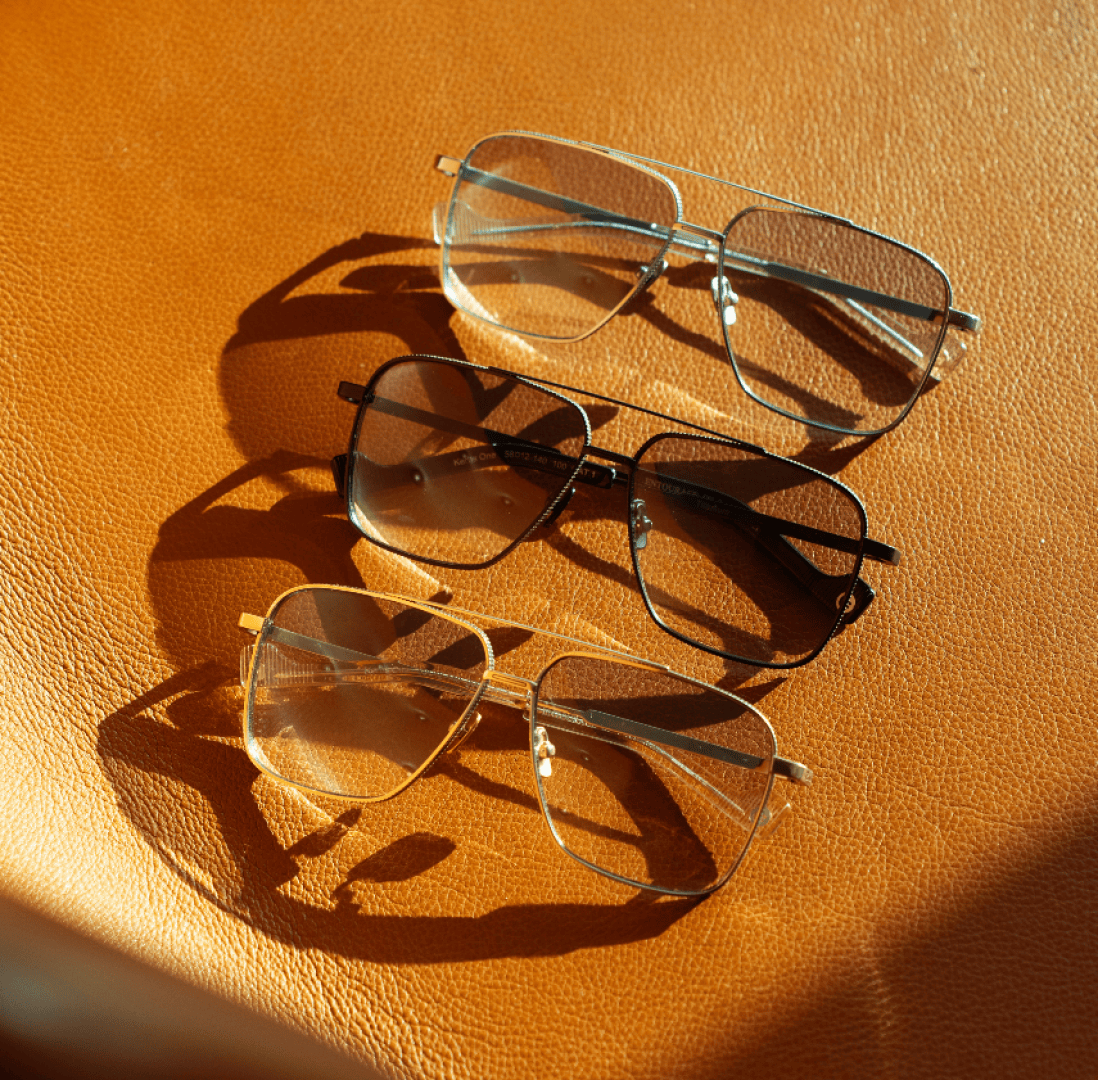 Drei Brillen im Pilotenstil mit goldenen, schwarzen und silbernen Rahmen sind in einer Reihe auf einer strukturierten braunen Lederoberfläche angeordnet und werfen im Sonnenlicht unterschiedliche Schatten.