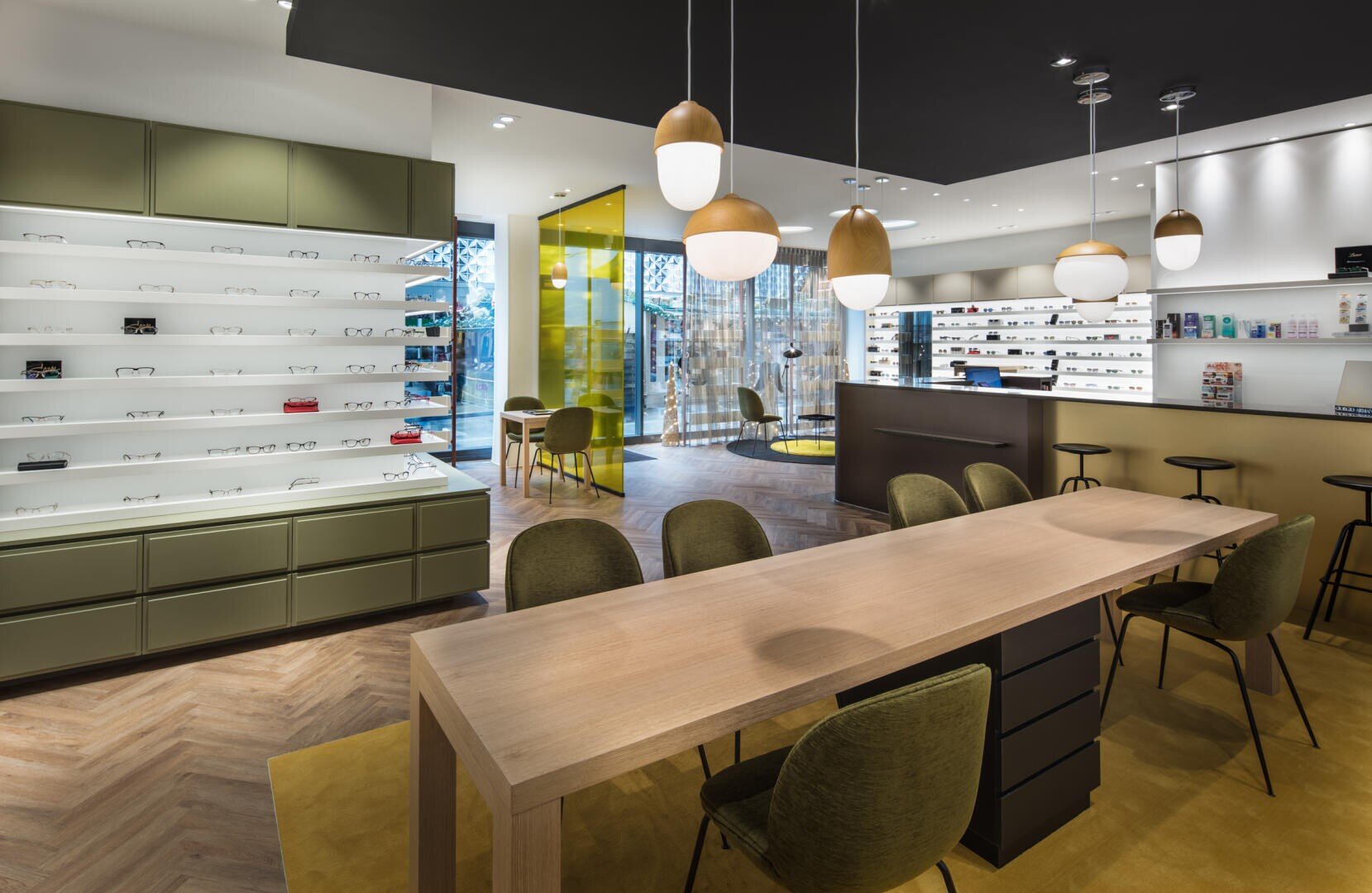 Modernes Optikergeschäft mit olivgrünen Regalen, einem Holztisch mit Stühlen, Pendelleuchten und an den Wänden ausgestellten Brillen. Der Raum hat ein modernes Design mit klaren Linien und warmer Beleuchtung.