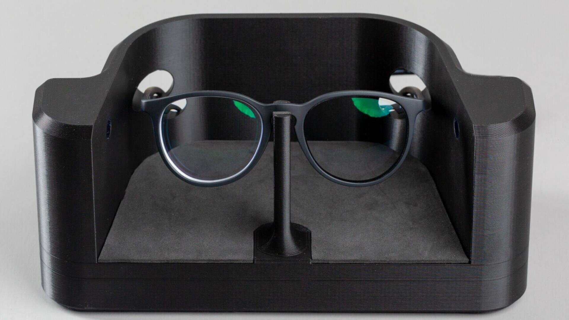 A pair of black-rimmed glasses is displayed on a stand inside a black, rectangular enclosure with curved edges and a soft gray base.