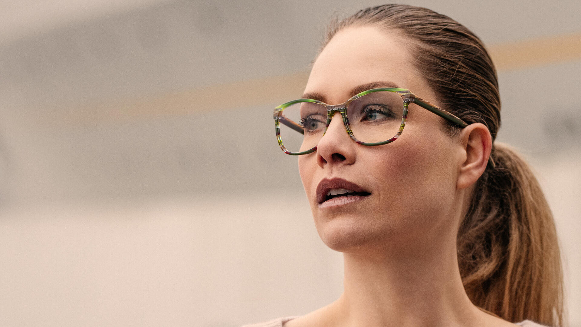 Eine Frau mit heller Haut und zurückgebundenem braunen Haar trägt eine bunte, gemusterte Brille und blickt nachdenklich zur Seite. Der Hintergrund ist unscharf und neutral.