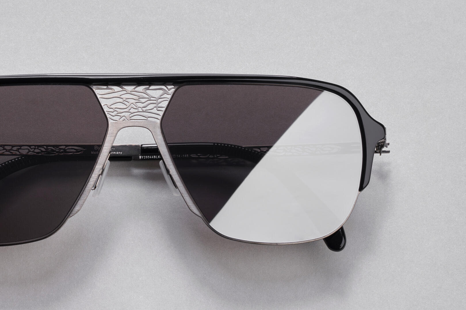 Nahaufnahme einer stilvollen Sonnenbrille mit silbernem Metallrahmen und dunklen Gläsern, die auf dem Nasensteg und dem oberen Rand vor einem hellgrauen Hintergrund ein kompliziertes Gravurmuster aufweist.