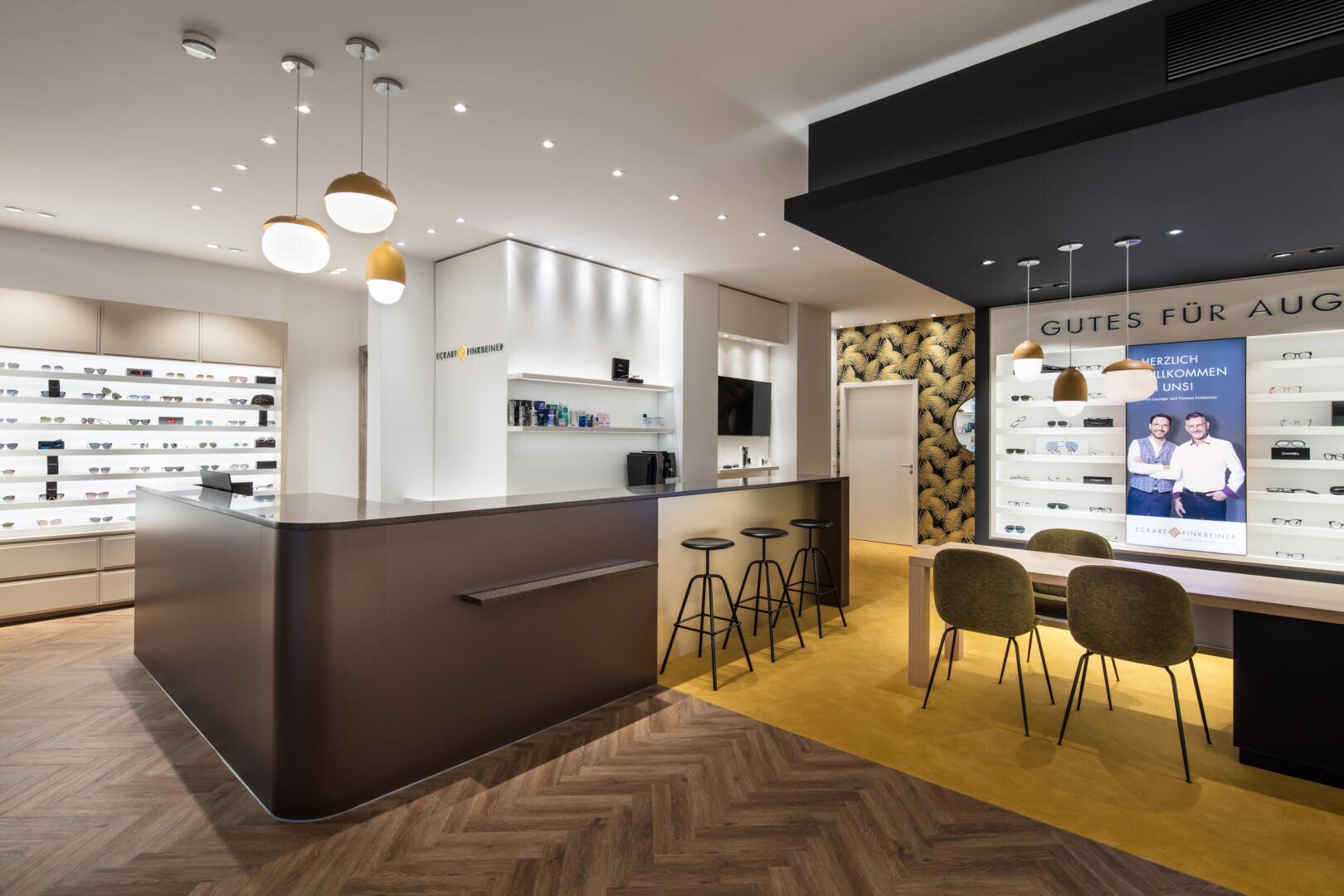 Modernes Optikergeschäft mit eleganten Theken, Barhockern, mit Brillen gefüllten Ausstellungsregalen, Hängeleuchten und einem Sitzbereich. Eine Wand zeigt ein Schild in deutscher Sprache und ein Werbebild mit zwei Personen.