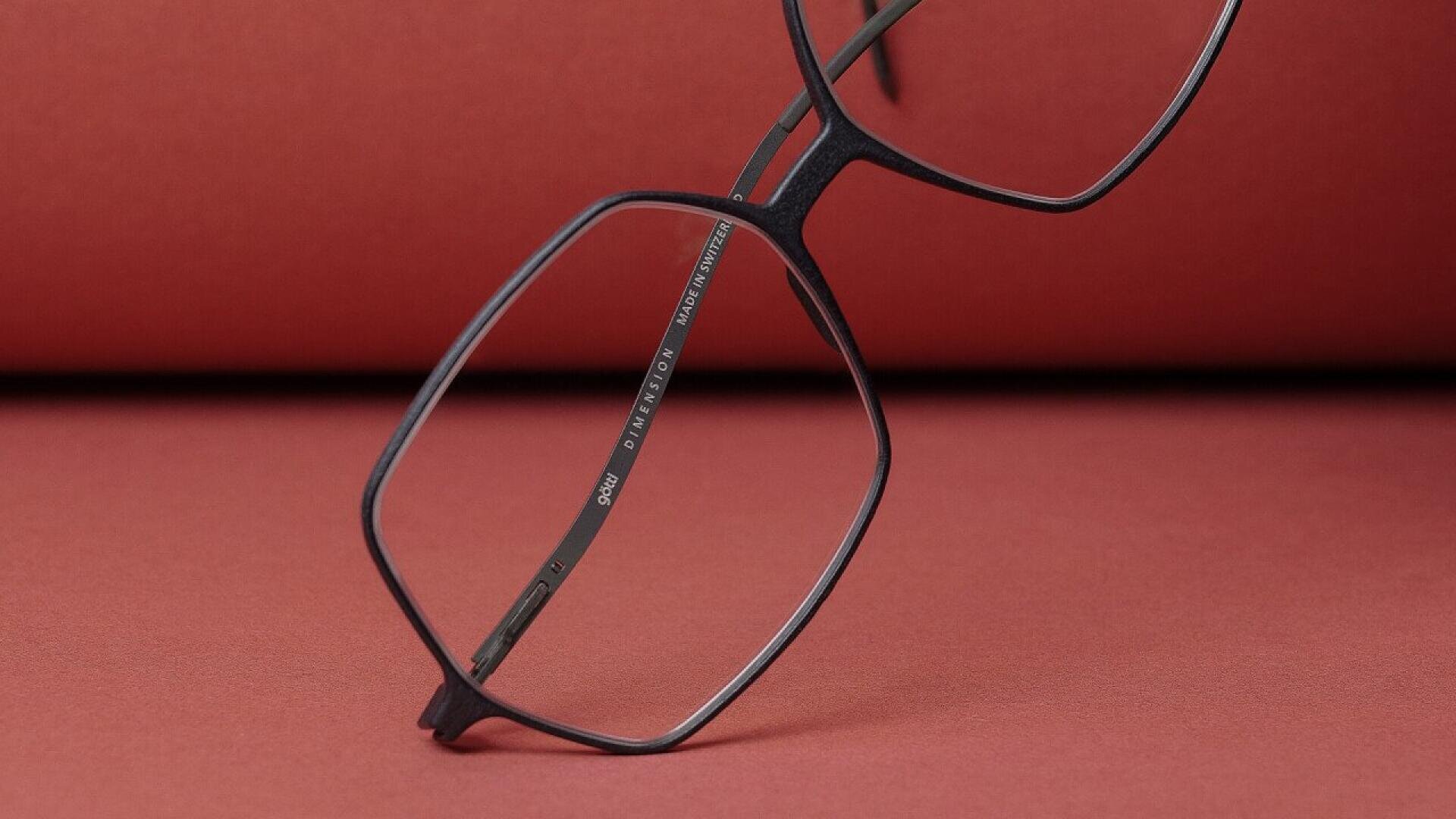 A pair of black, geometric eyeglasses with large lenses rests on a red surface, with a red background behind it. The image focuses on the details of the glasses frame.