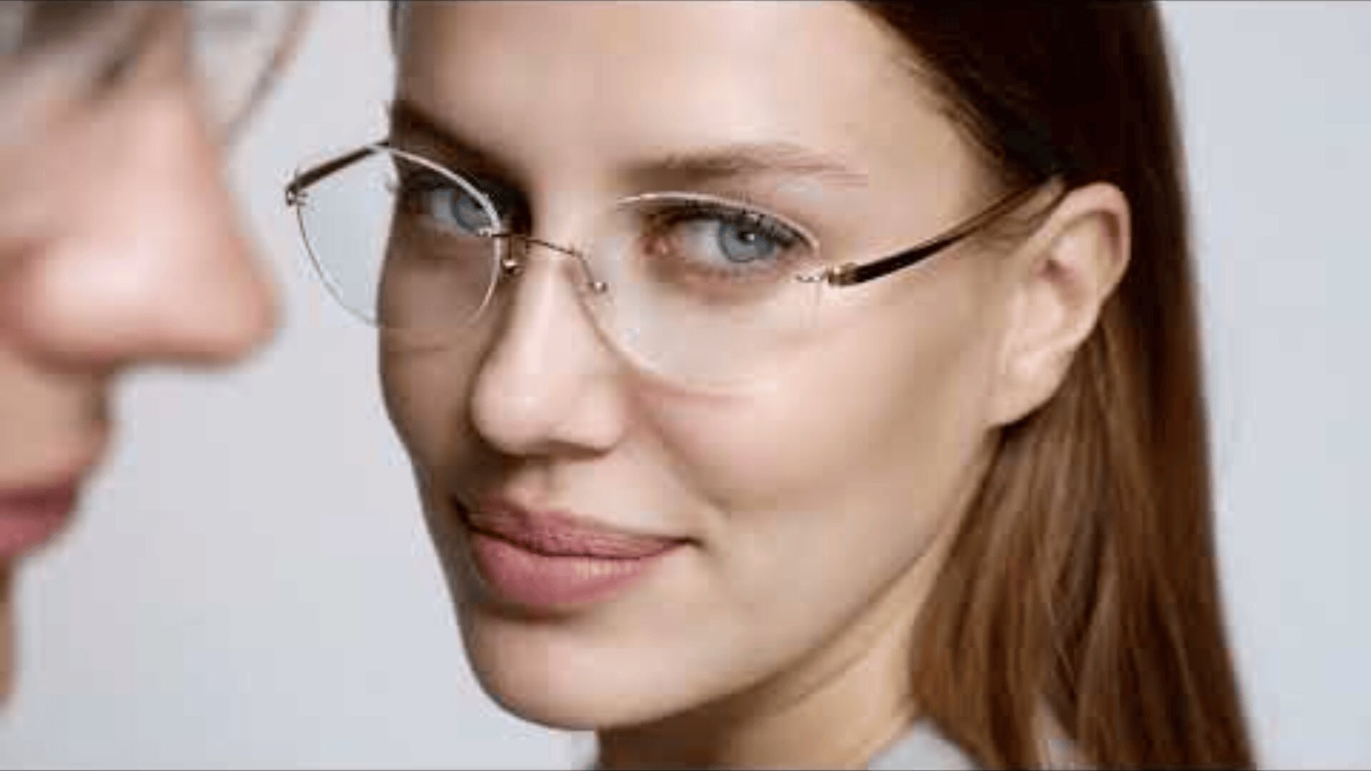 Eine Frau mit glattem braunem Haar und klarer Brille blickt mit einem leichten Lächeln in die Kamera. Auf der linken Seite des Bildes ist eine weitere Person zu sehen, die teilweise unscharf ist.