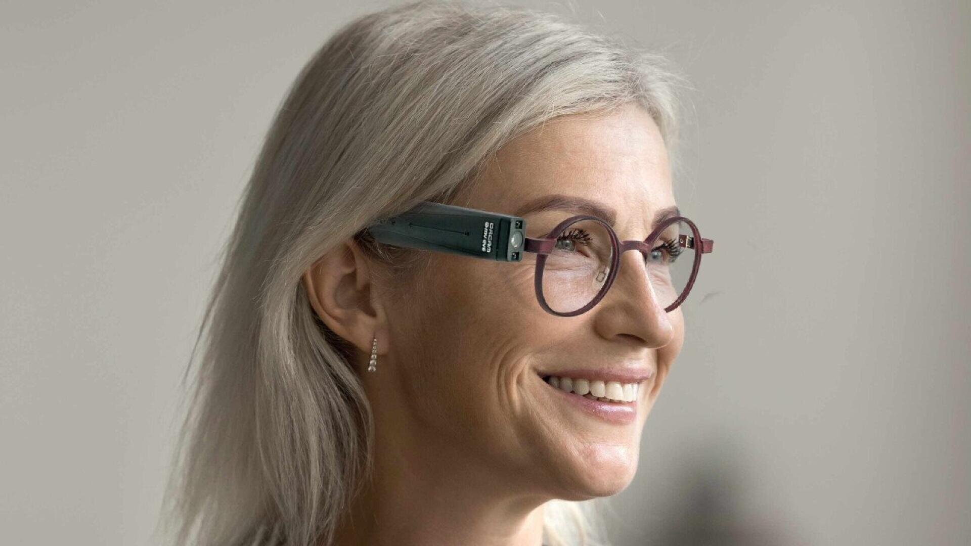 A woman with long gray hair and round eyeglasses is smiling. She is wearing a small hearing aid behind her ear and pearl earrings. The background is blurred and neutral.