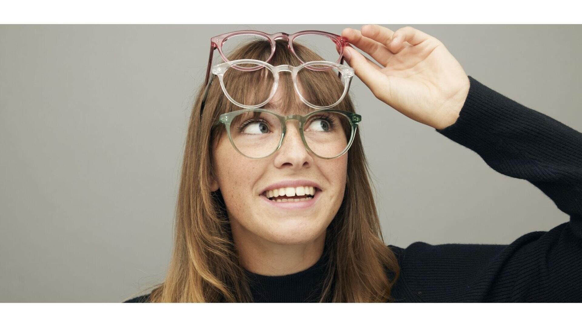 A smiling woman wearing green glasses holds a second pair of pink glasses above her head, looking upward. She has long brown hair and is dressed in a dark sweater against a plain background.