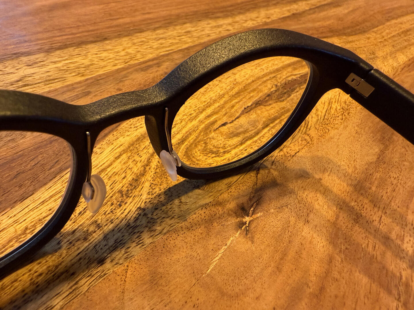 Black-rimmed eyeglasses with nose pads resting on a wooden surface, with warm lighting highlighting the wood grain.