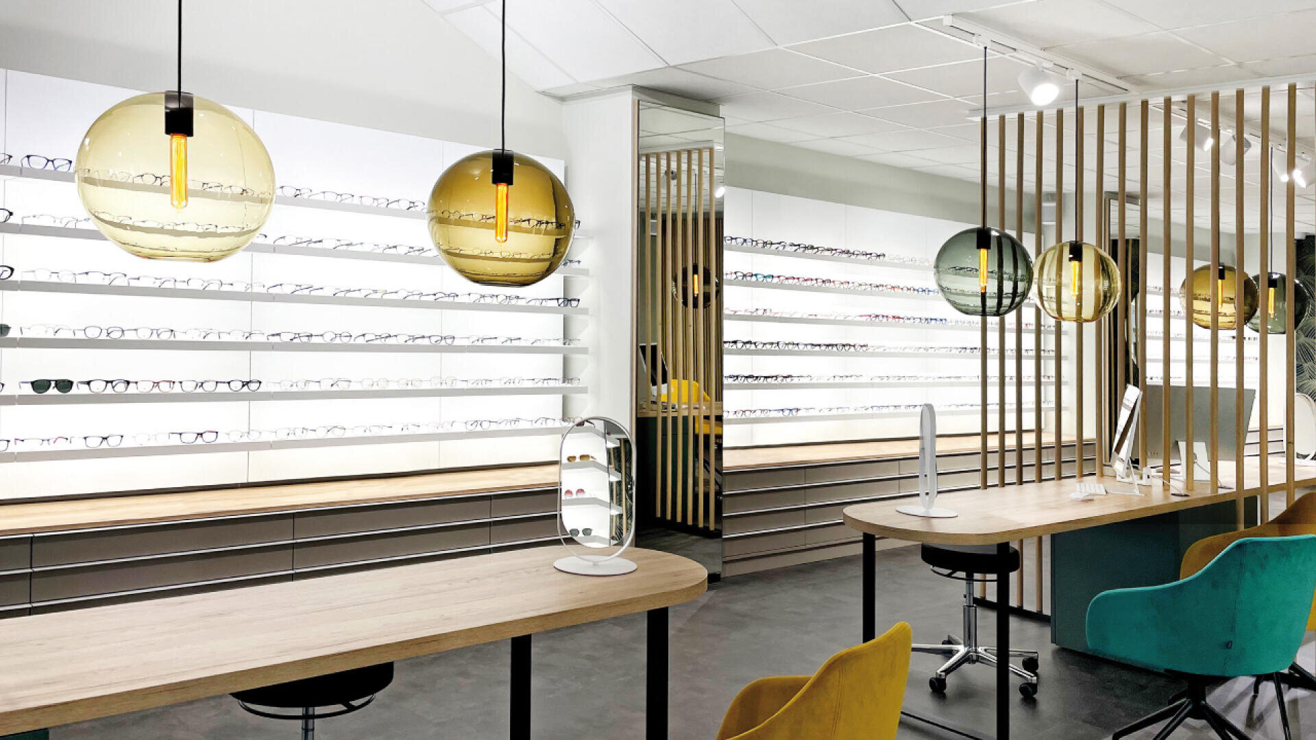 Modern optician store interior with rows of eyeglasses displayed on white shelves, wooden tables with chairs, colorful glass pendant lights, and a computer on the desk. The space is bright and stylish.