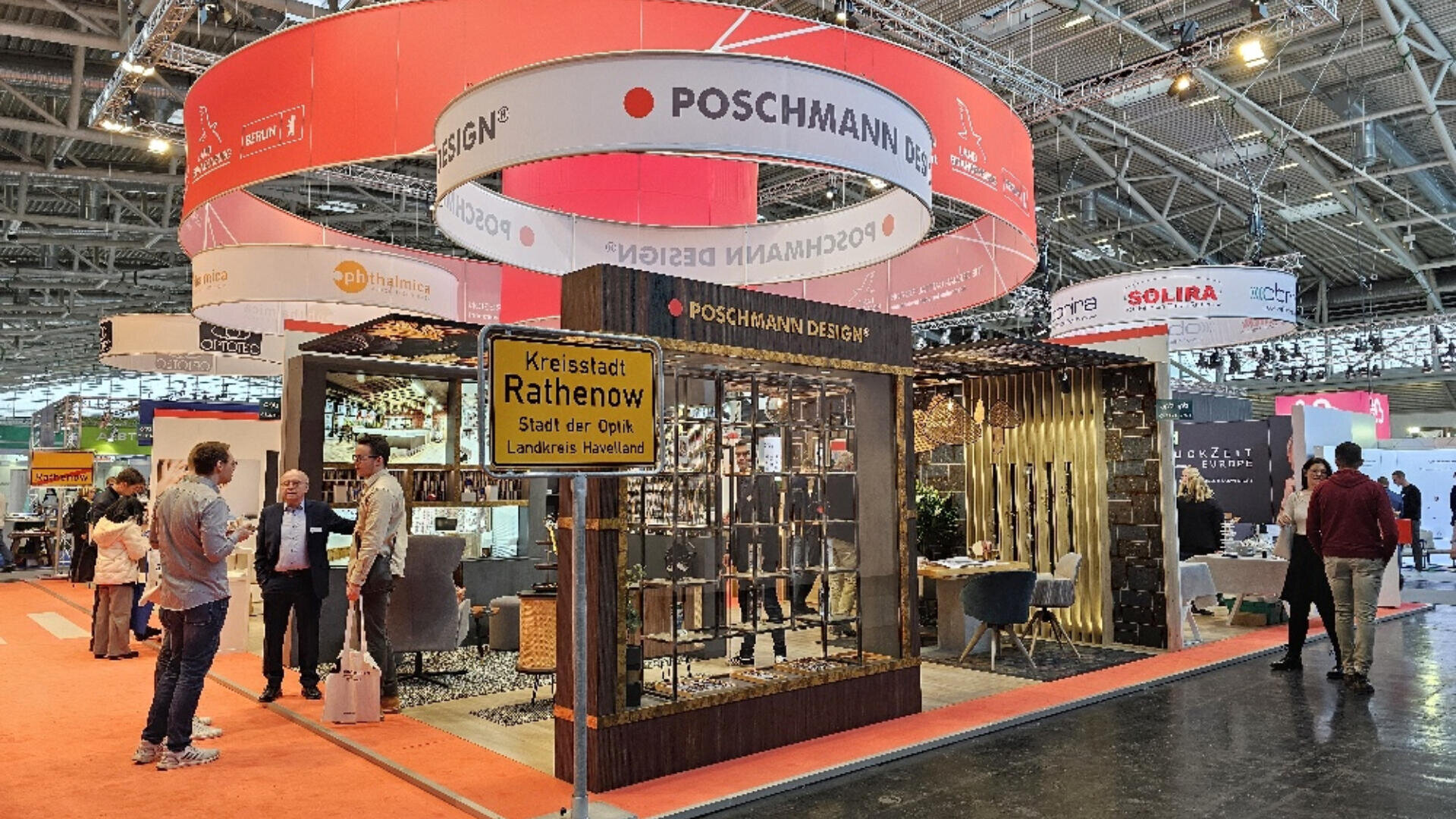 A trade show booth for POSCHMANN DESIGN features modern furniture and decor, with several people talking nearby. Overhead, large circular banners hang under a steel and glass ceiling.