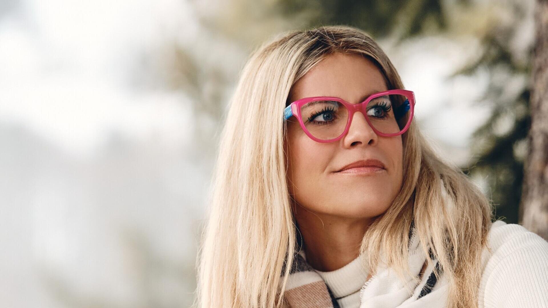 A woman with long blonde hair and bright pink glasses looks to the side outdoors. She is wearing a white turtleneck sweater and a scarf, and trees are blurred in the background.