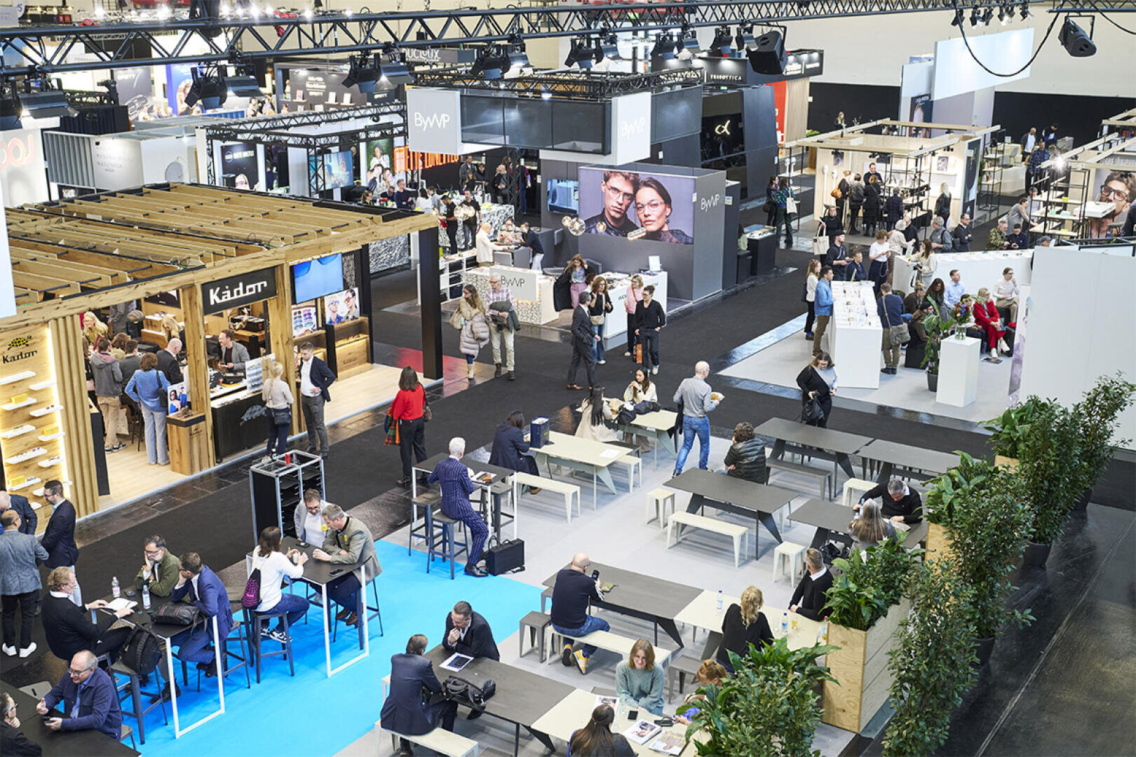 Eine belebte Messe oder Ausstellungshalle mit verschiedenen Ständen, Displays und Menschen, die herumlaufen, Kontakte knüpfen und an Tischen sitzen. Die Szene ist lebendig mit moderner Dekoration, Pflanzen und Bannern, die für Produkte und Marken werben.