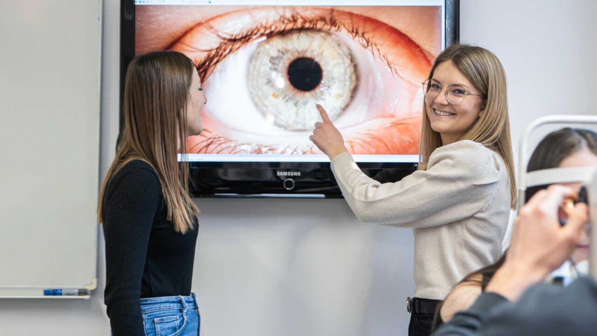 Two women stand in front of a screen displaying a close-up image of an eye; one woman points at the screen while smiling, and the other faces her. A third person is partially visible in the foreground.