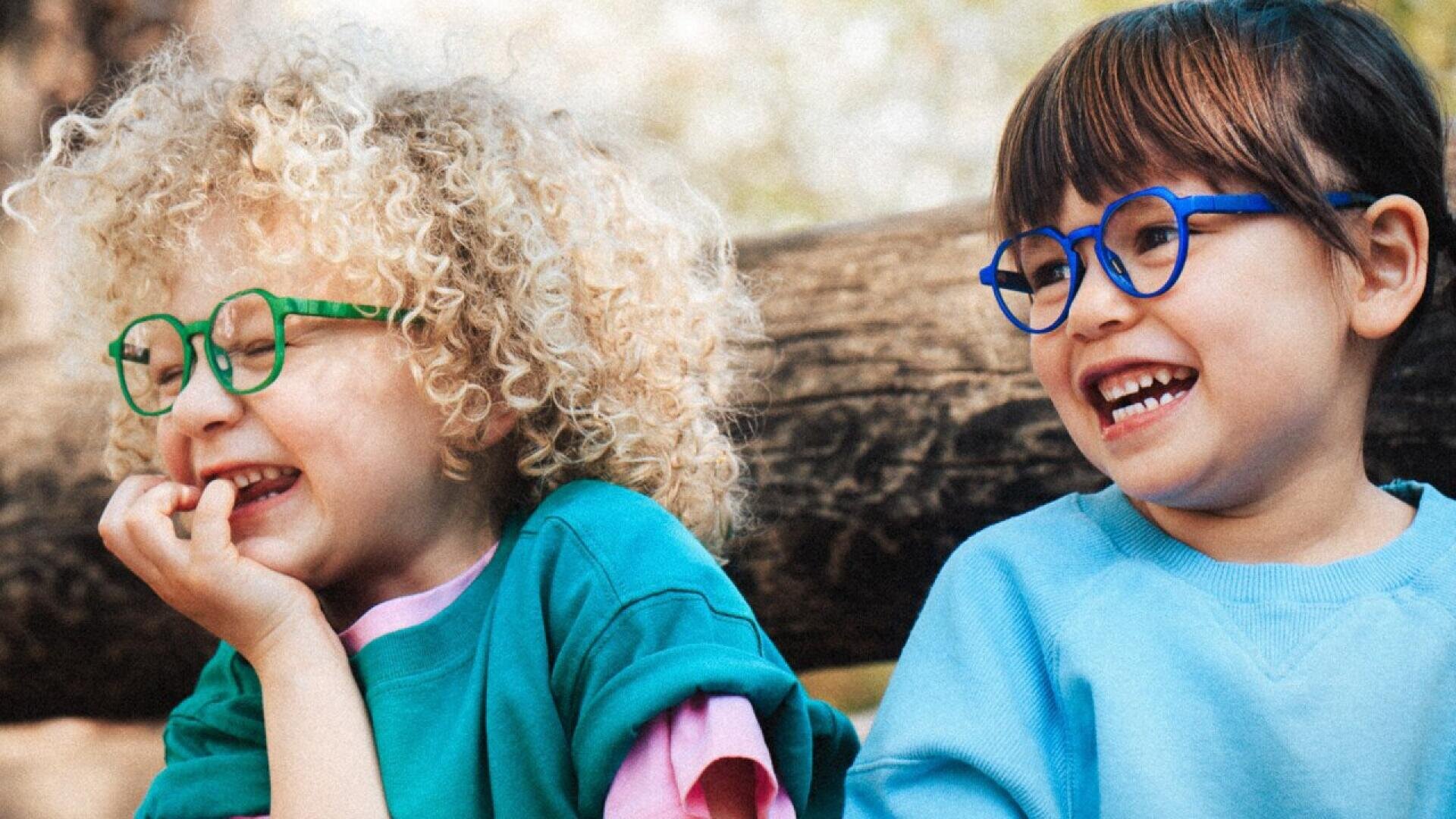 Two young children with curly and straight hair, both wearing colorful glasses and light blue shirts, sit close together outdoors, smiling and laughing in front of a blurred tree trunk background.