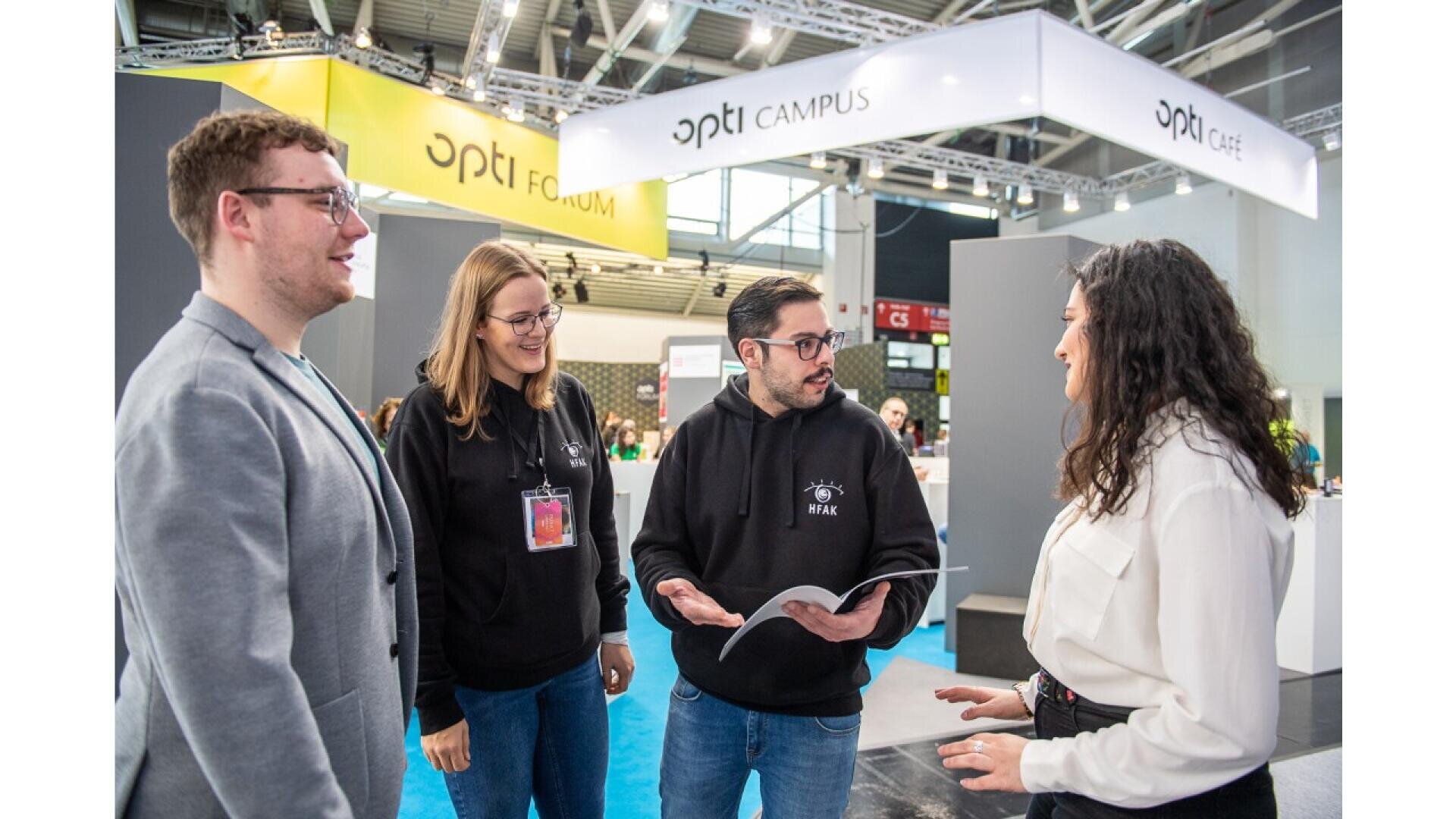 Four people stand and talk at an indoor event or expo, with two wearing black hoodies, one in a grey blazer, and one in a white shirt. Bright signs above read “opti FORUM,” “opti CAMPUS,” and “opti CAFE.”.