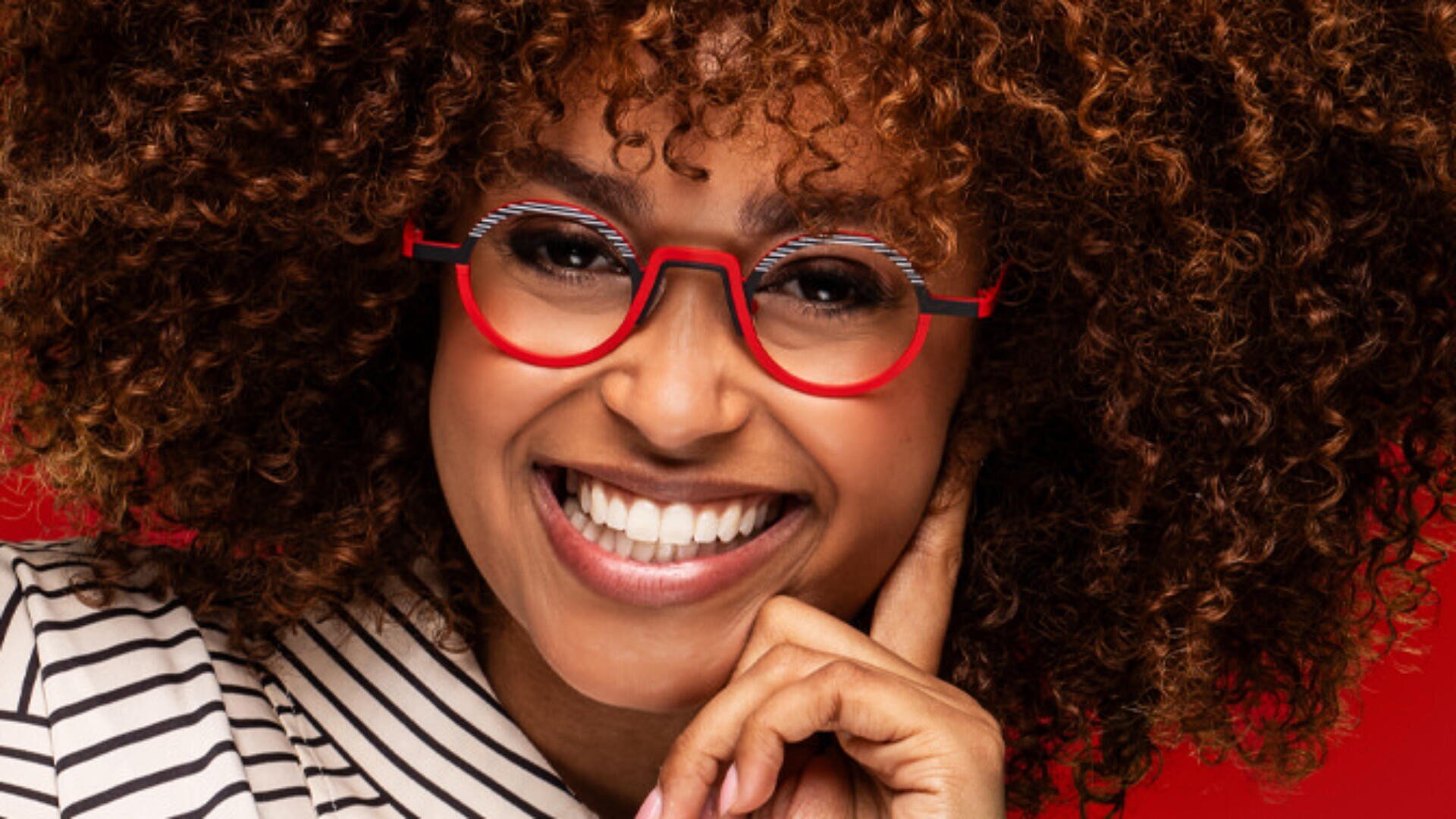 A smiling person with curly brown hair wears red glasses and a striped shirt, resting their chin on their hand against a red background.