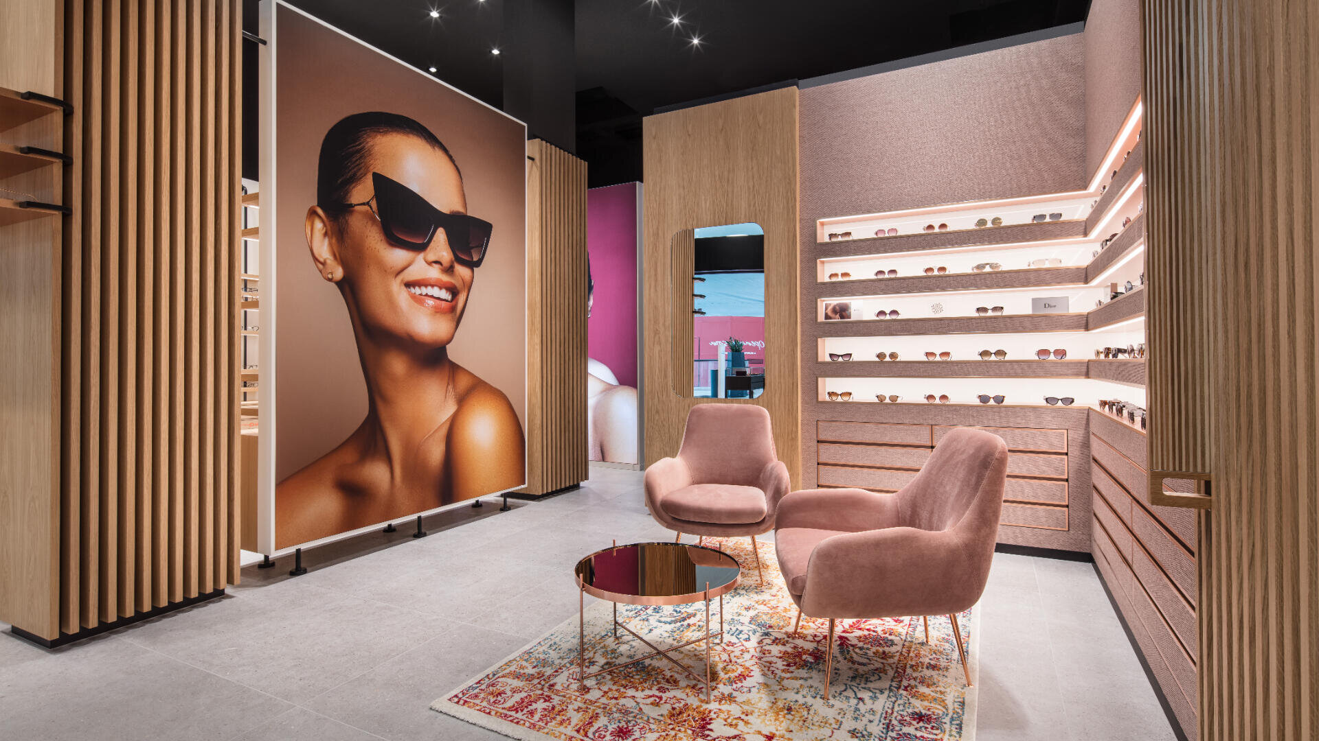 Modernes Interieur eines Brillengeschäfts mit rosa Stühlen, einem kleinen runden Tisch auf einem gemusterten Teppich, hölzernen Ausstellungsregalen mit Sonnenbrillen und einem großen Poster einer lächelnden Frau mit Sonnenbrille.