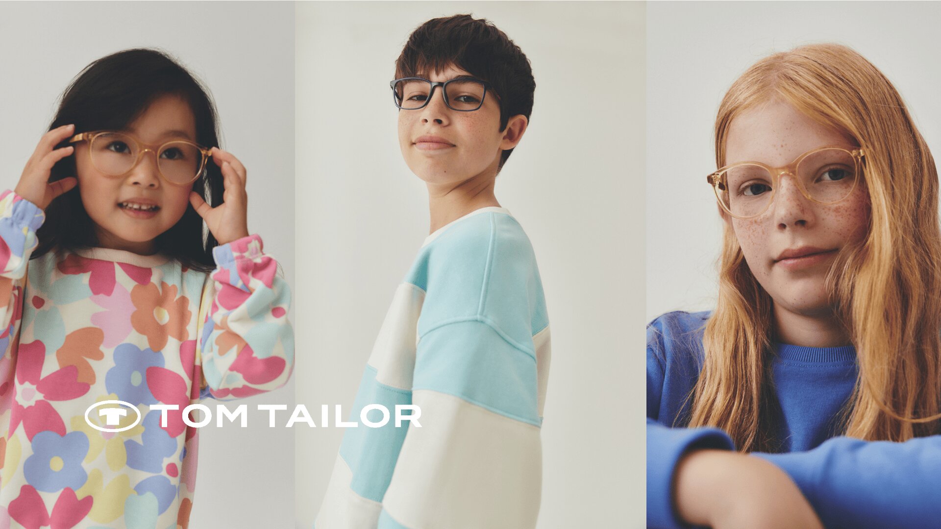 Drei Kinder mit Brillen und bunten, lässigen Pullovern stehen vor einem hellen Hintergrund. Das Tom Tailor-Logo ist in der unteren linken Ecke des Bildes zu sehen.