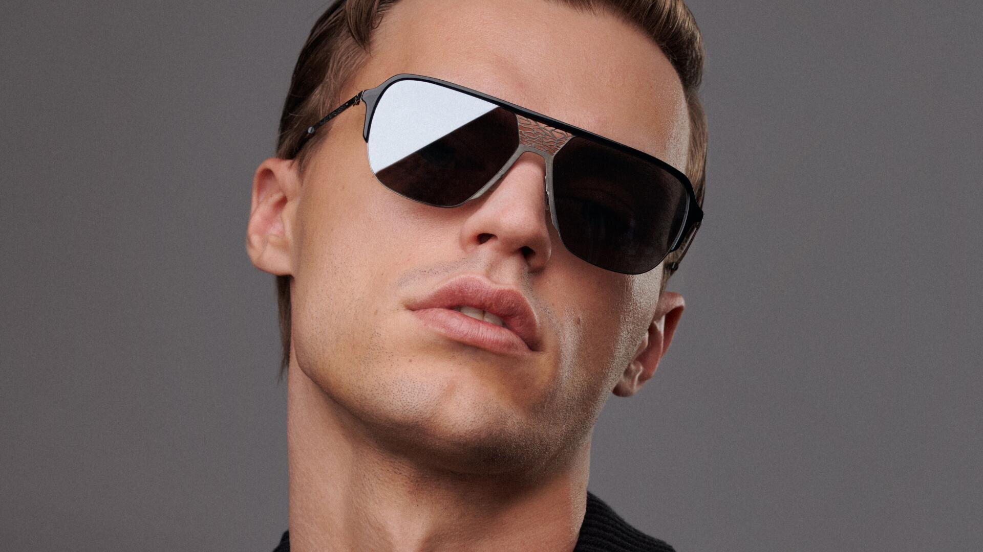 Ein junger Mann mit einer großen, dunklen Sonnenbrille blickt selbstbewusst in die Kamera. Er hat helle Haut, kurzes braunes Haar und einen neutralen Gesichtsausdruck vor einem schlichten grauen Hintergrund.