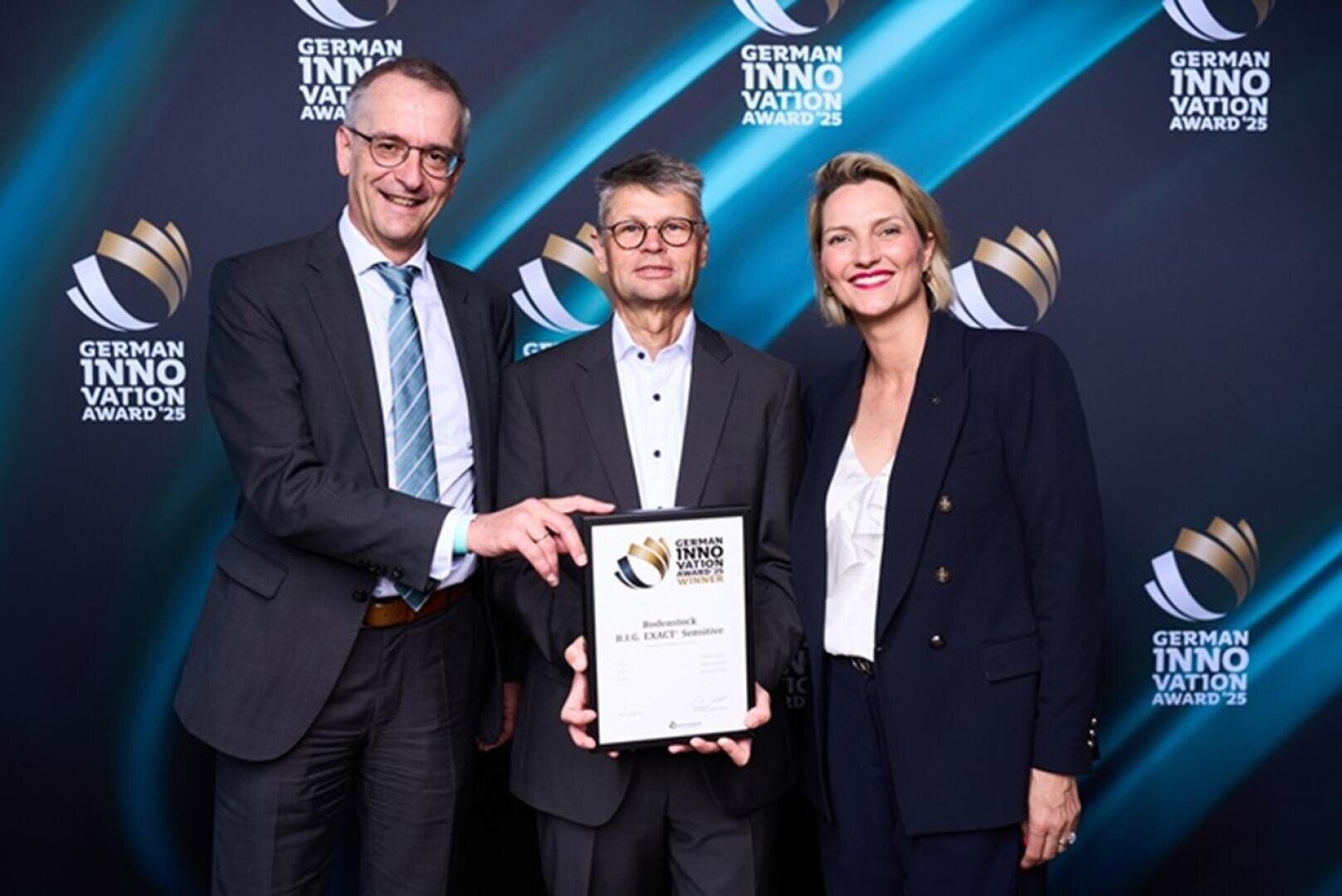 Drei Personen in Businesskleidung stehen lächelnd mit einer gerahmten Urkunde bei der Verleihung des German Innovation Award vor einem schwarzen Hintergrund, auf dem immer wieder der Name und das Logo der Veranstaltung zu sehen sind.