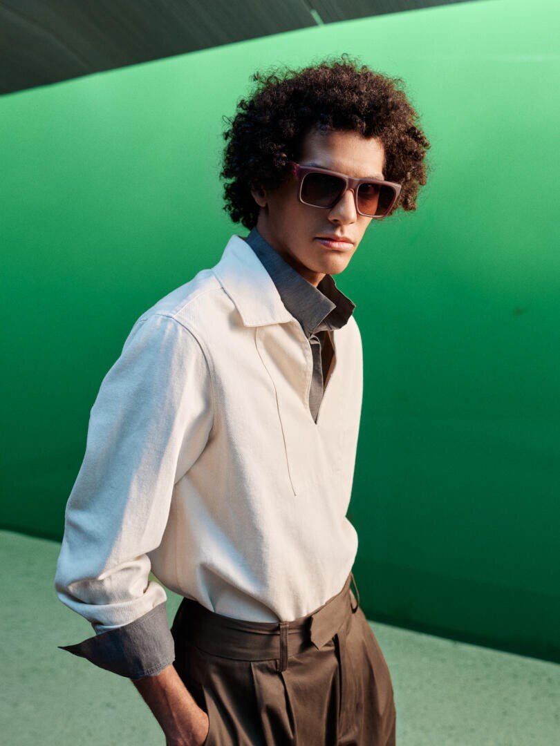 Ein Mann mit lockigem Haar, der eine Sonnenbrille, ein weißes Hemd mit grauem Kragen und eine hochgeschnittene Bundfaltenhose trägt, steht vor einem hellgrünen Hintergrund.