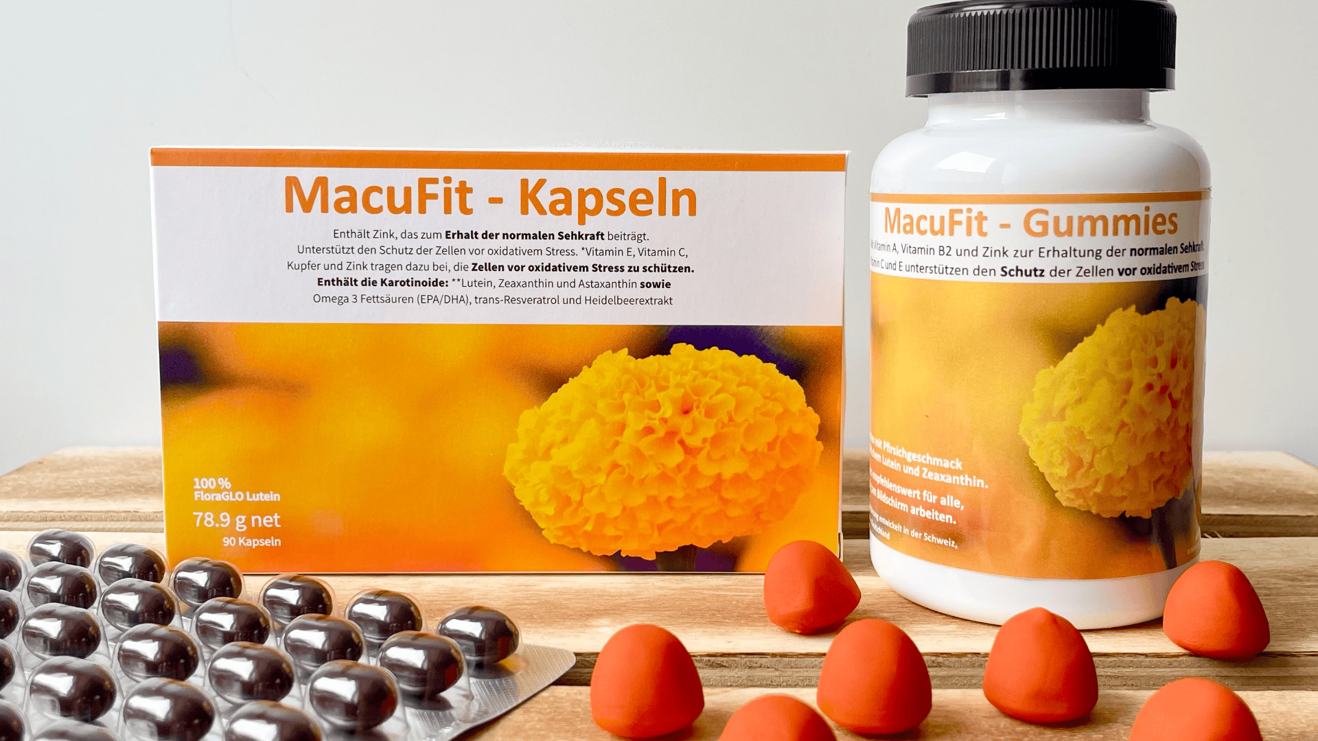 Auf einem Tisch stehen eine Schachtel und eine Flasche mit der Aufschrift MacuFit in deutscher Sprache und einer orangefarbenen Verpackung, daneben liegen eine Blisterpackung mit Kapseln und mehrere orangefarbene, herzförmige Gummibärchen.