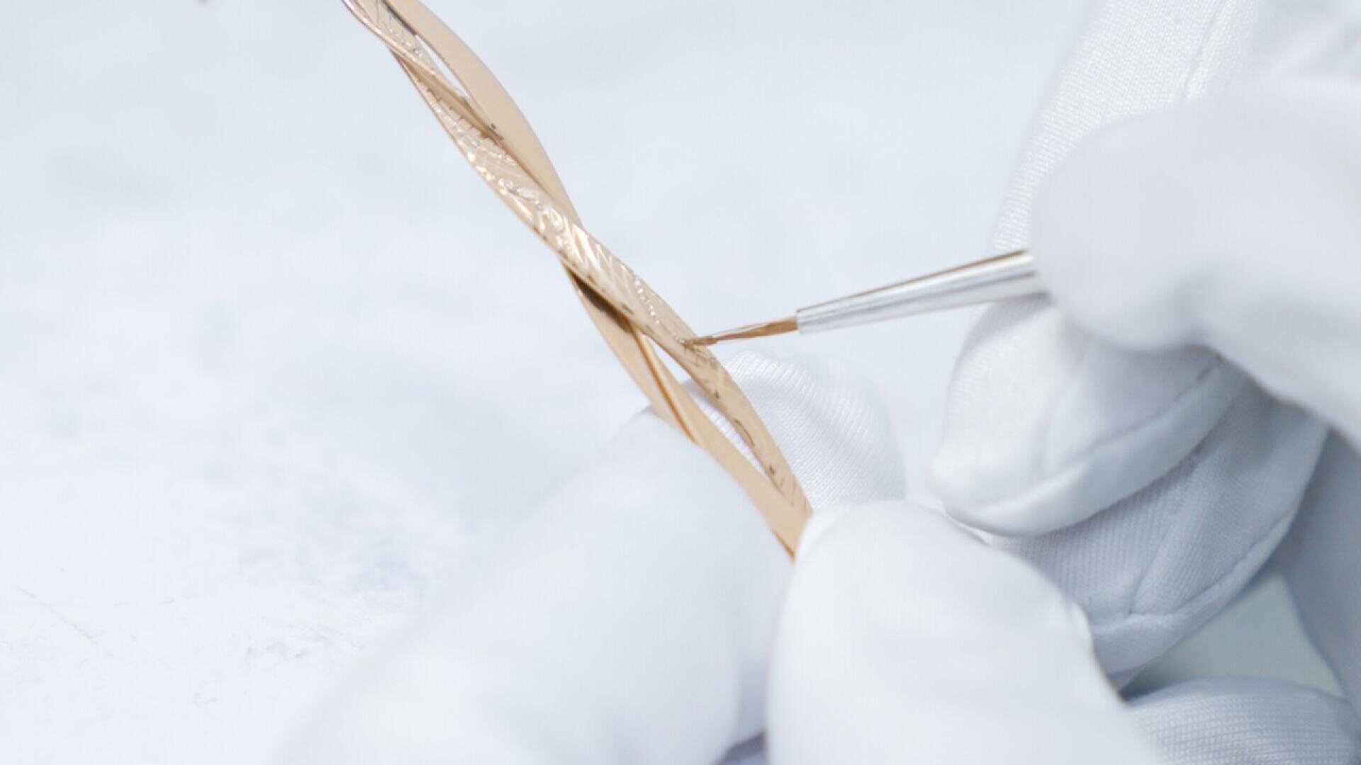 A gloved hand uses a small brush to carefully paint or apply detail to a twisted piece of gold jewelry against a white background.